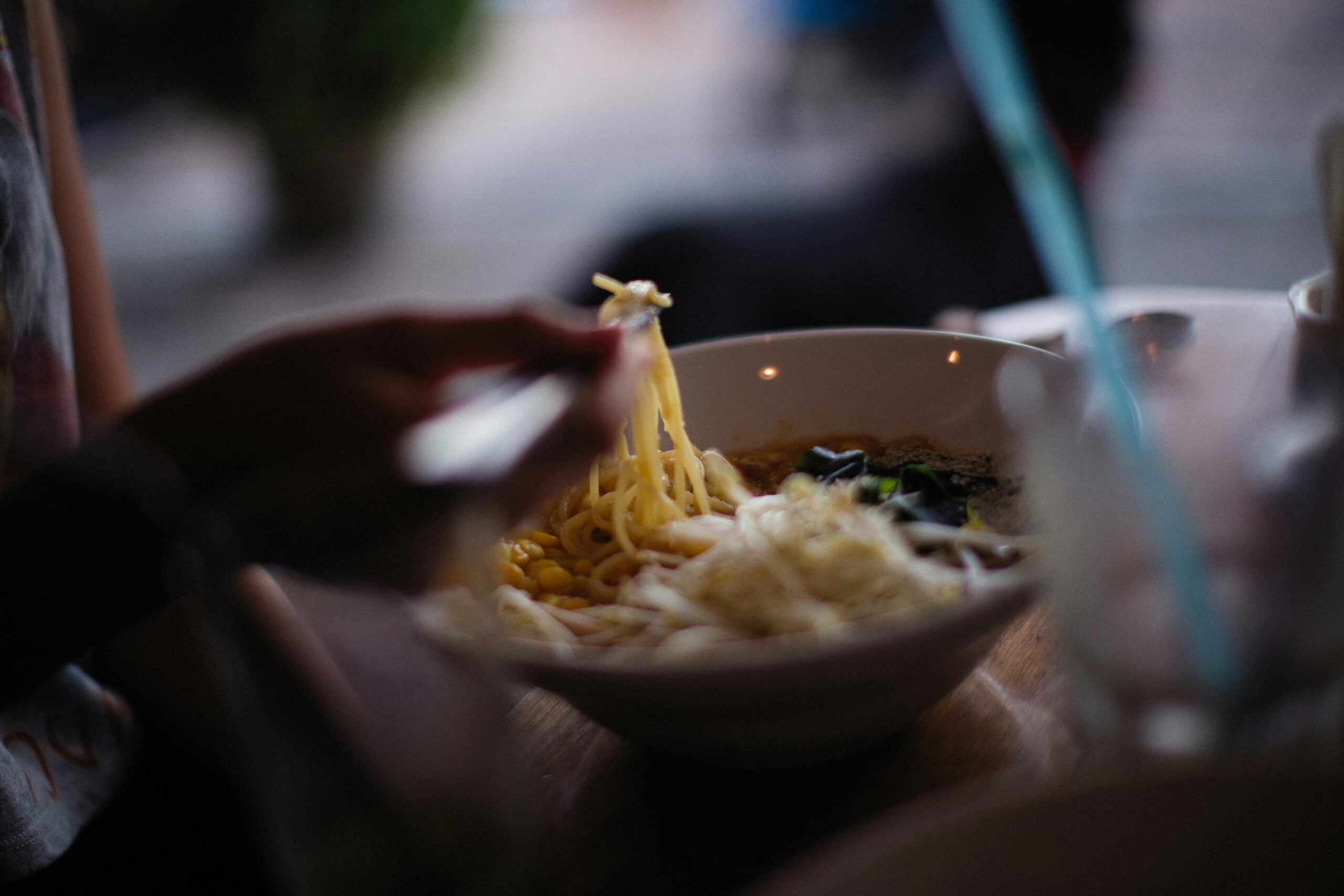 A close-up of a person using chopsticks to lift noodles from a bowl of soup, with various ingredients visible in the bowl on a wooden table. The background is softly blurred.