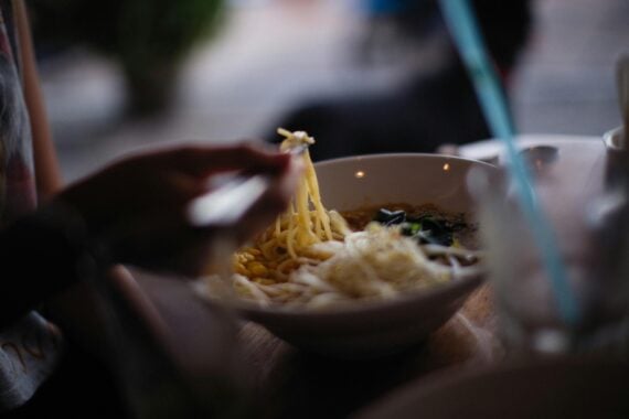 A person holding chopsticks lifts noodles from a bowl of soup, with another bowl and drinking glass visible on a wooden table in a softly lit setting.