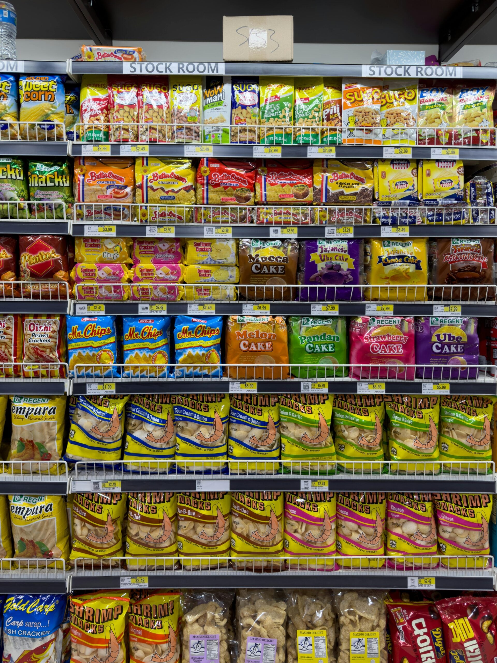 Shelves in a grocery store are filled with various brightly colored packaged snacks, cakes, and chips. A brown box labeled "SBK" sits on the top shelf, which is marked "STOCK ROOM.