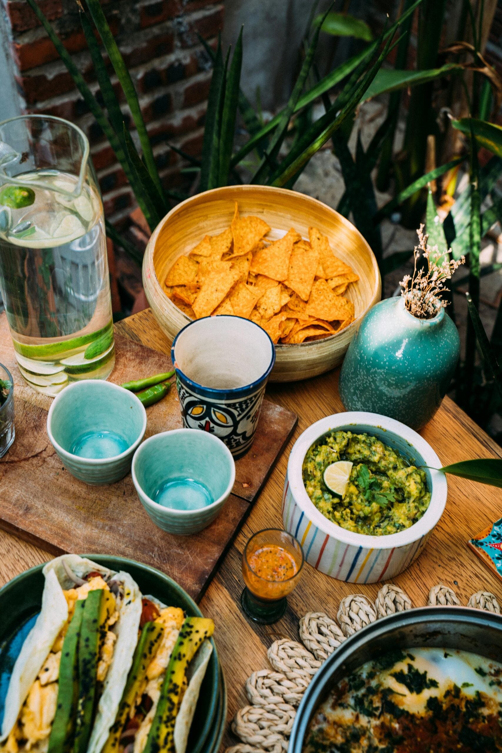 A wooden table set with tortilla chips, guacamole with lime, tacos, a small bottle of hot sauce, three ceramic cups, a glass of water with lime, and a green vase with dried flowers. Lush plants are in the background.