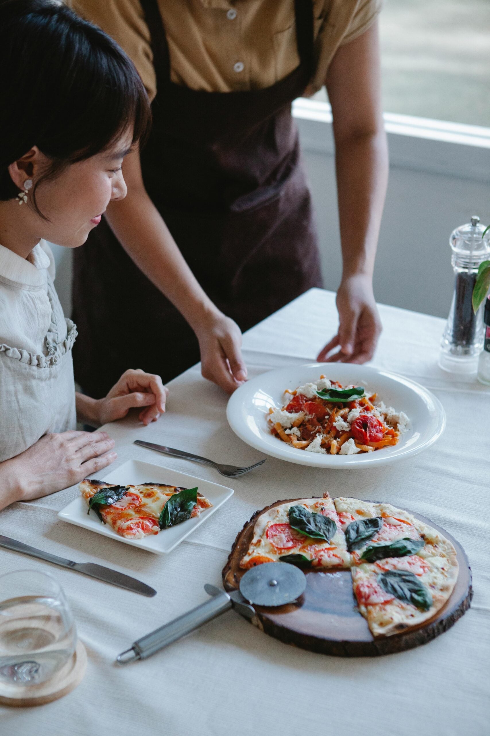 A woman sits at a table with a slice of pizza on her plate as another person serves a pasta dish. A pizza with basil leaves is on the table, along with cutlery, a glass, and a pepper grinder.