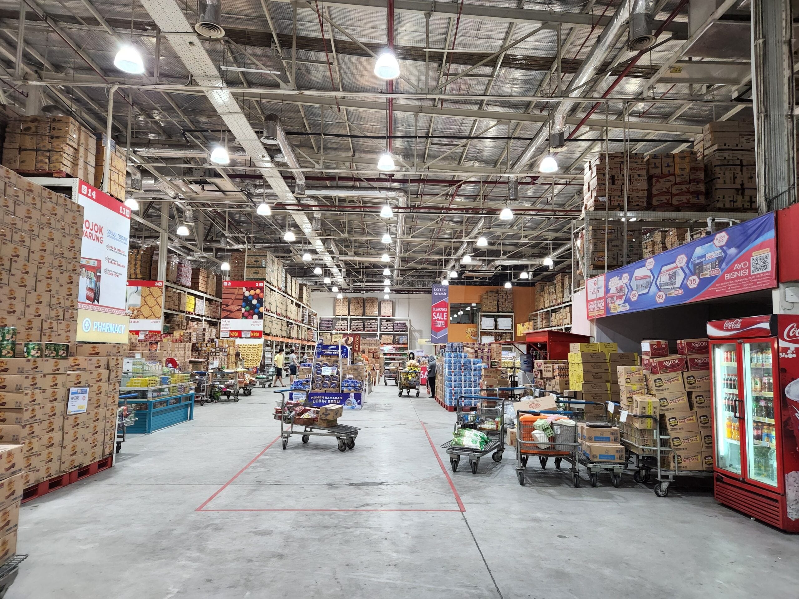 A wholesale warehouse with high shelves stacked with boxes, shopping carts filled with bulk goods, and a few visible refrigerators and freezers along the right side under bright overhead lighting.