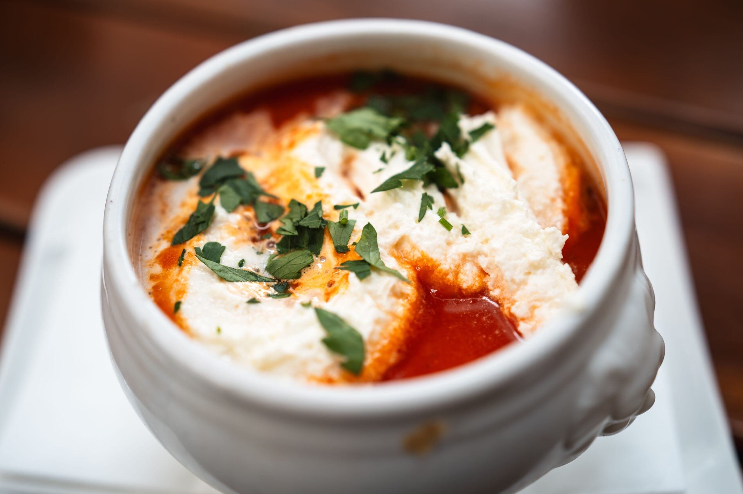 A close-up of a white bowl filled with tomato soup, topped with dollops of cream and garnished with freshly chopped parsley. The bowl is placed on a white plate.