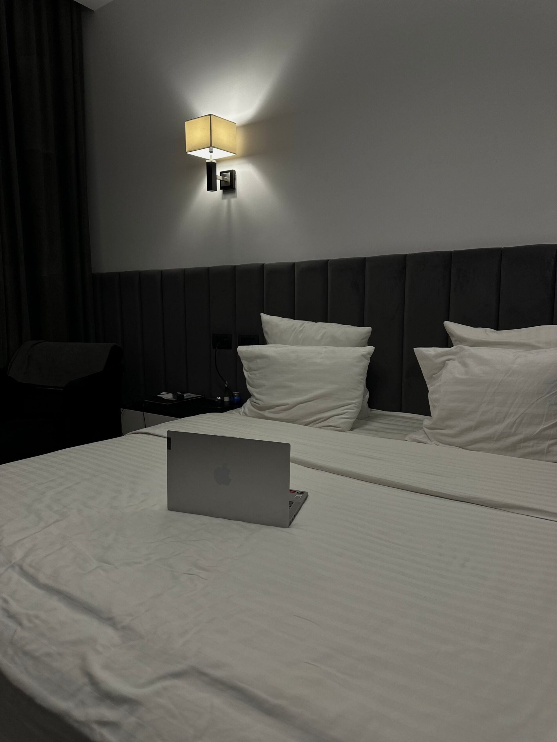 A silver laptop sits open on a neatly made bed with white linens, highlighting hotel room safety. A modern wall lamp illuminates the gray upholstered headboard while a dark curtain hangs to the left.