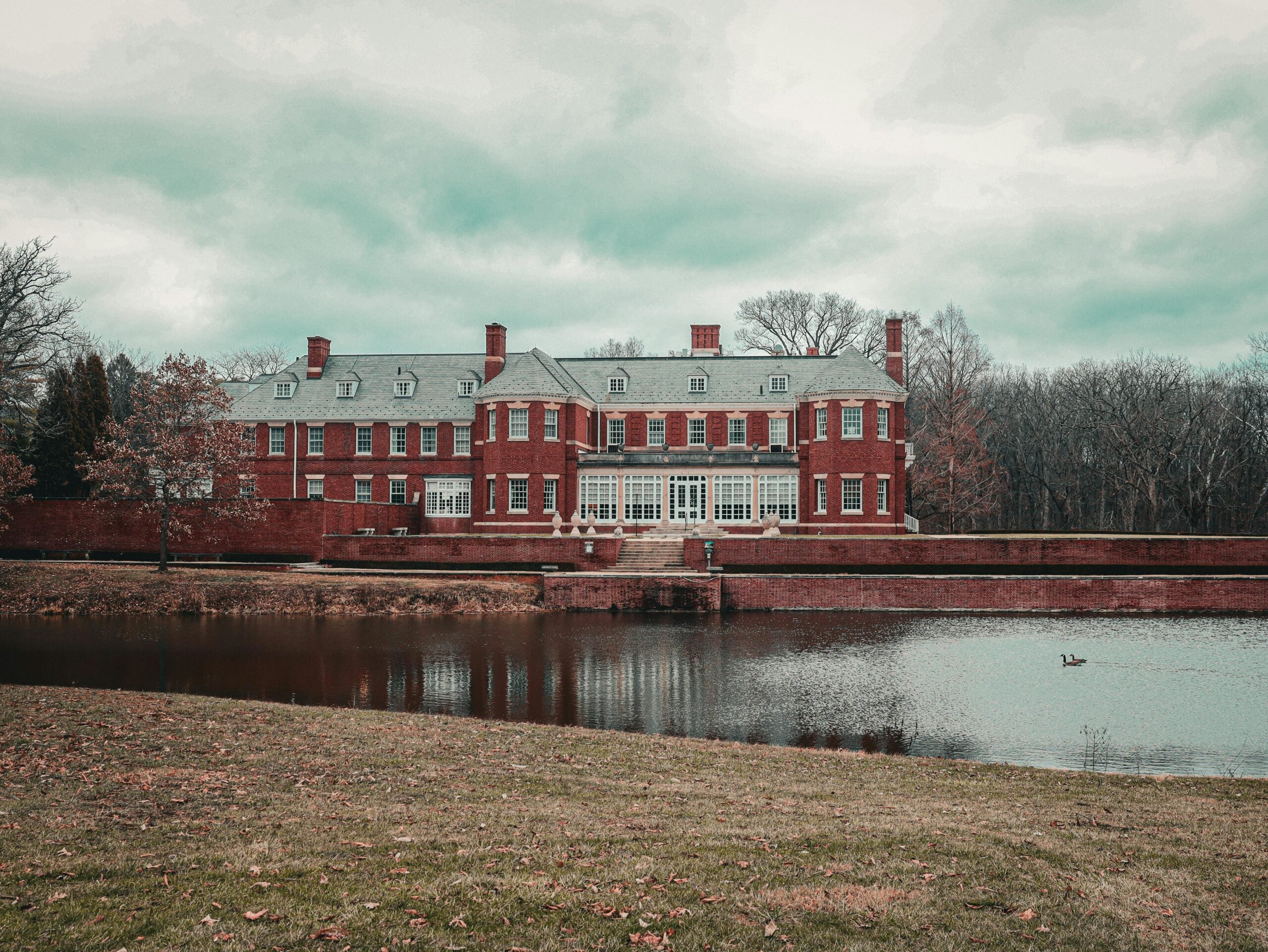 A large, red-brick mansion with many windows and chimneys sits behind a pond on a cloudy day, surrounded by leafless trees and a grassy lawn with scattered autumn leaves.
