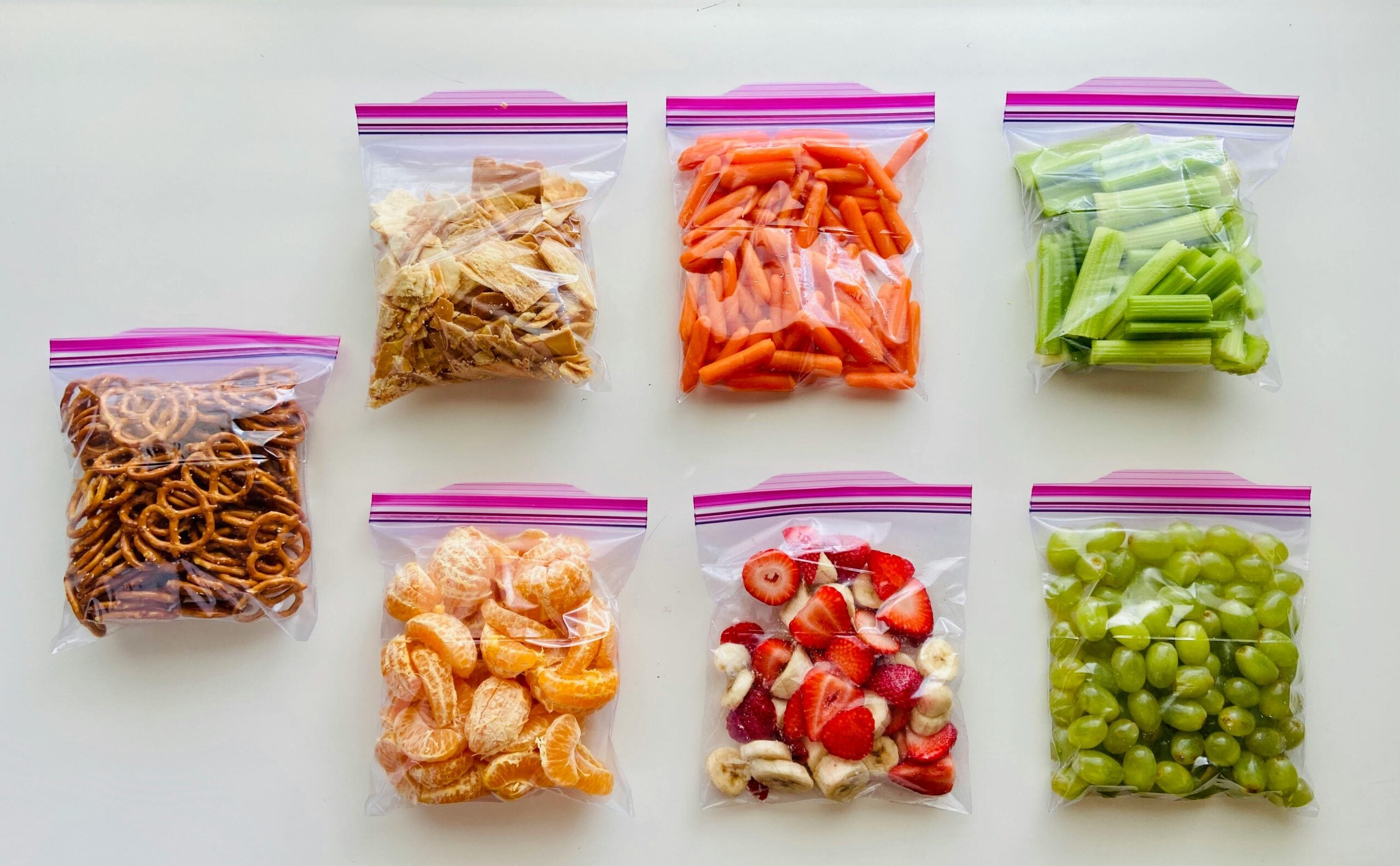 Seven clear ziplock bags containing snacks: top row has chips, carrot sticks, and celery sticks; bottom row has pretzels, orange slices, mixed strawberries and bananas, and green grapes, all neatly arranged on a white surface.