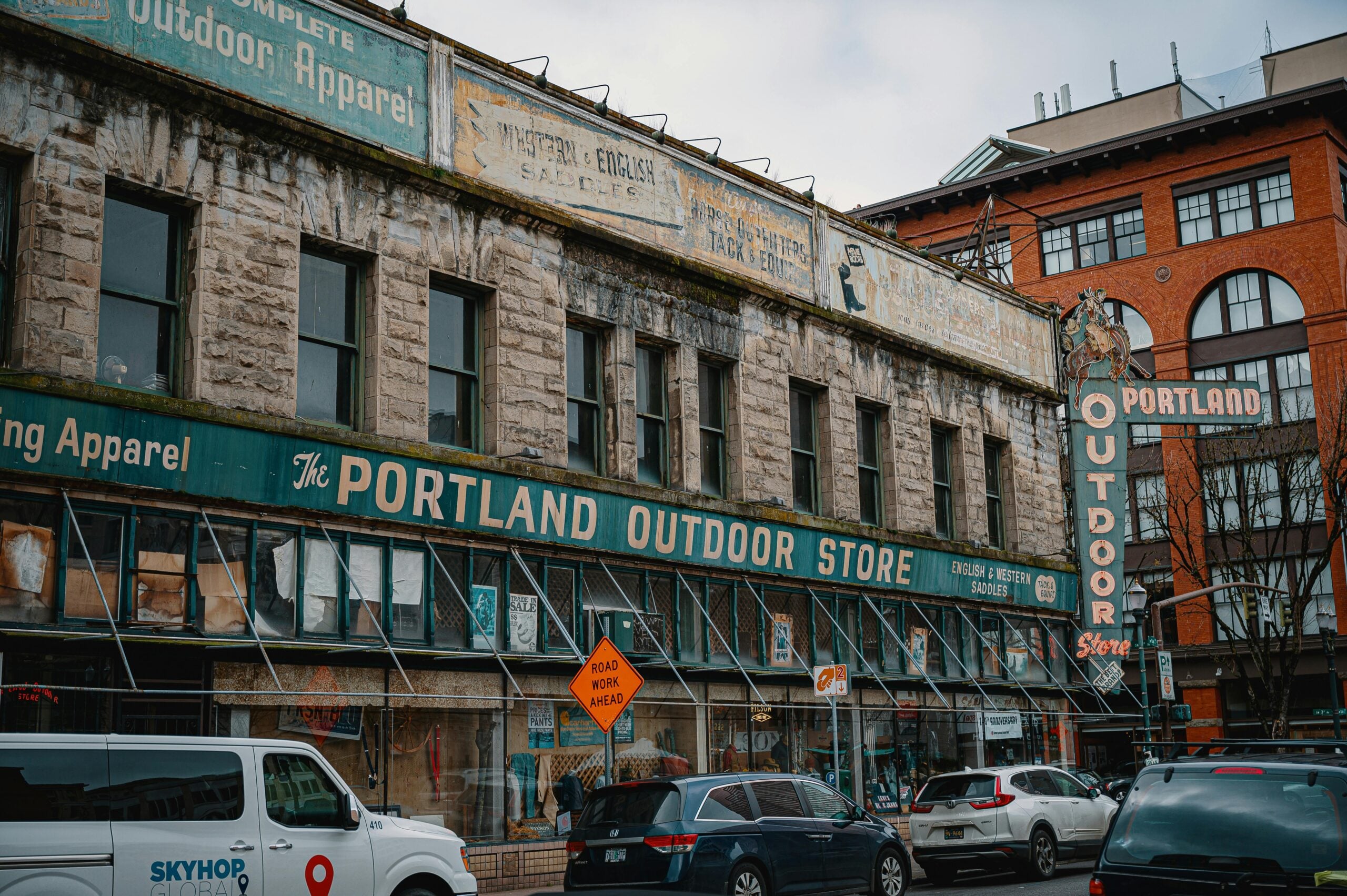 A street view of the Portland Outdoor Store with vintage signage on an old stone building. Several parked cars line the street in front of the store, and a cloudy sky is visible.