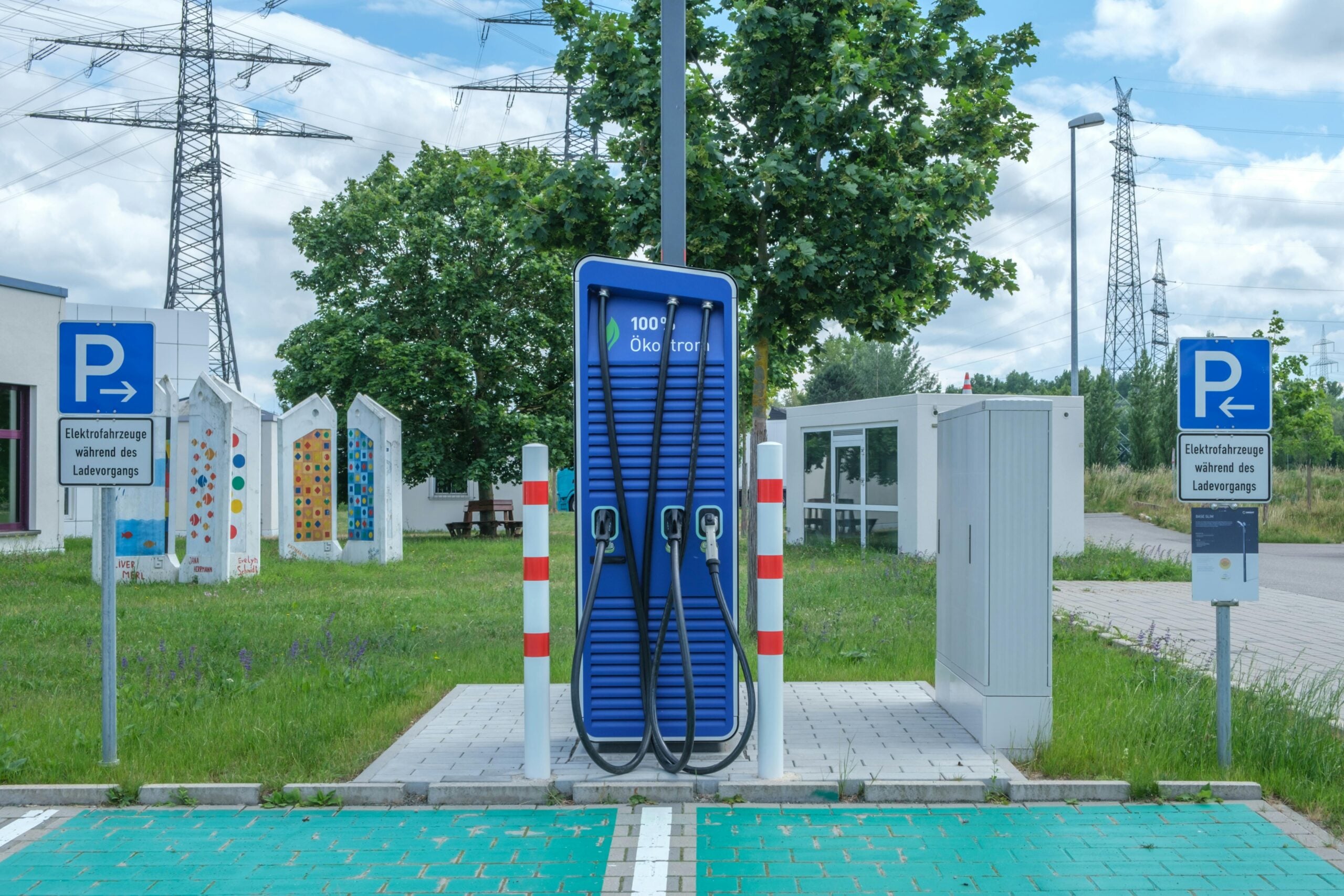 A blue electric vehicle charging station stands between two parking spaces marked for electric cars, with signs in German and trees, grass, and power lines visible in the background.