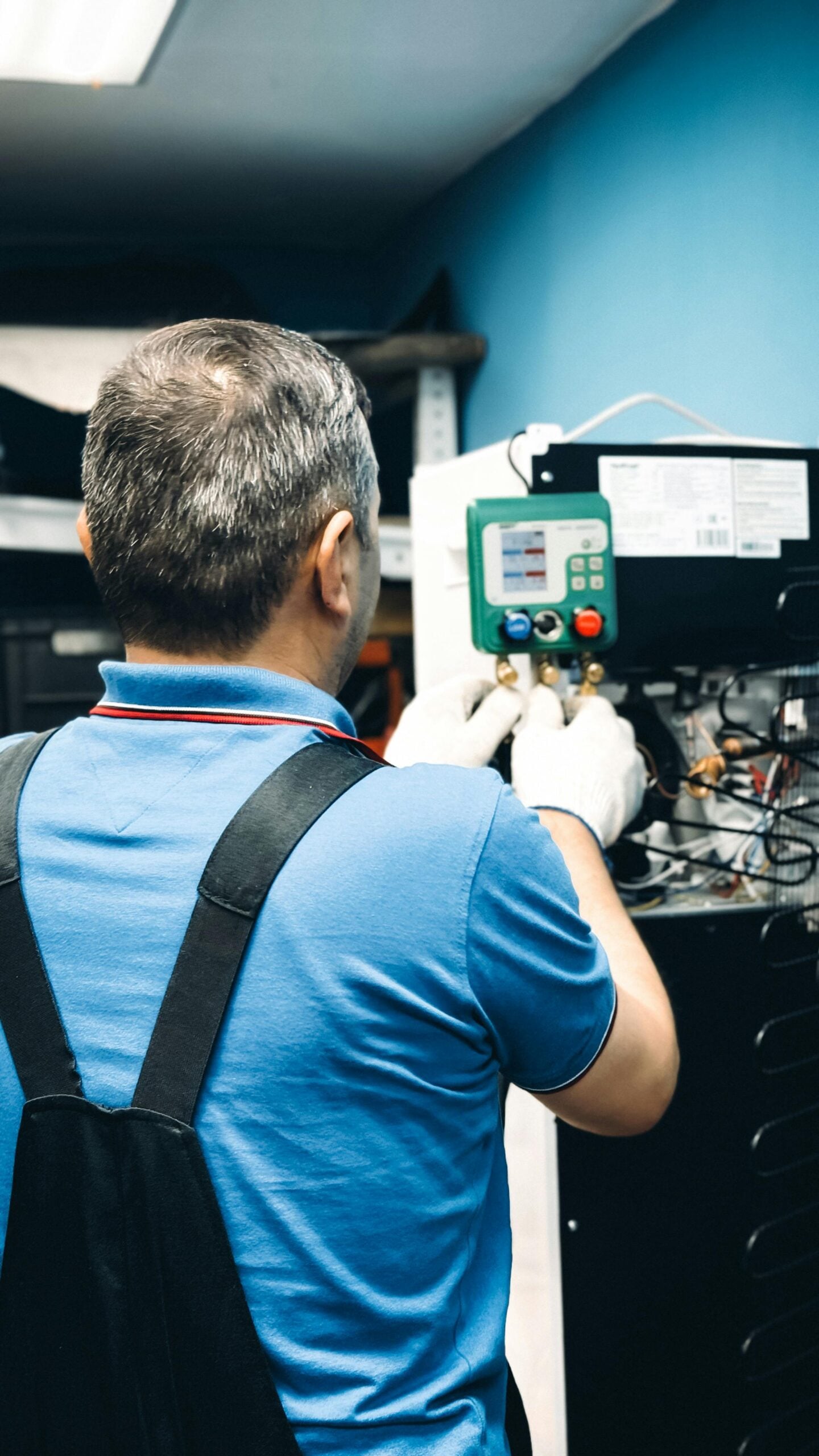 A technician in a blue shirt and black overalls uses a gauge to check equipment inside a machine, possibly performing maintenance or repairs in a workshop with blue walls.