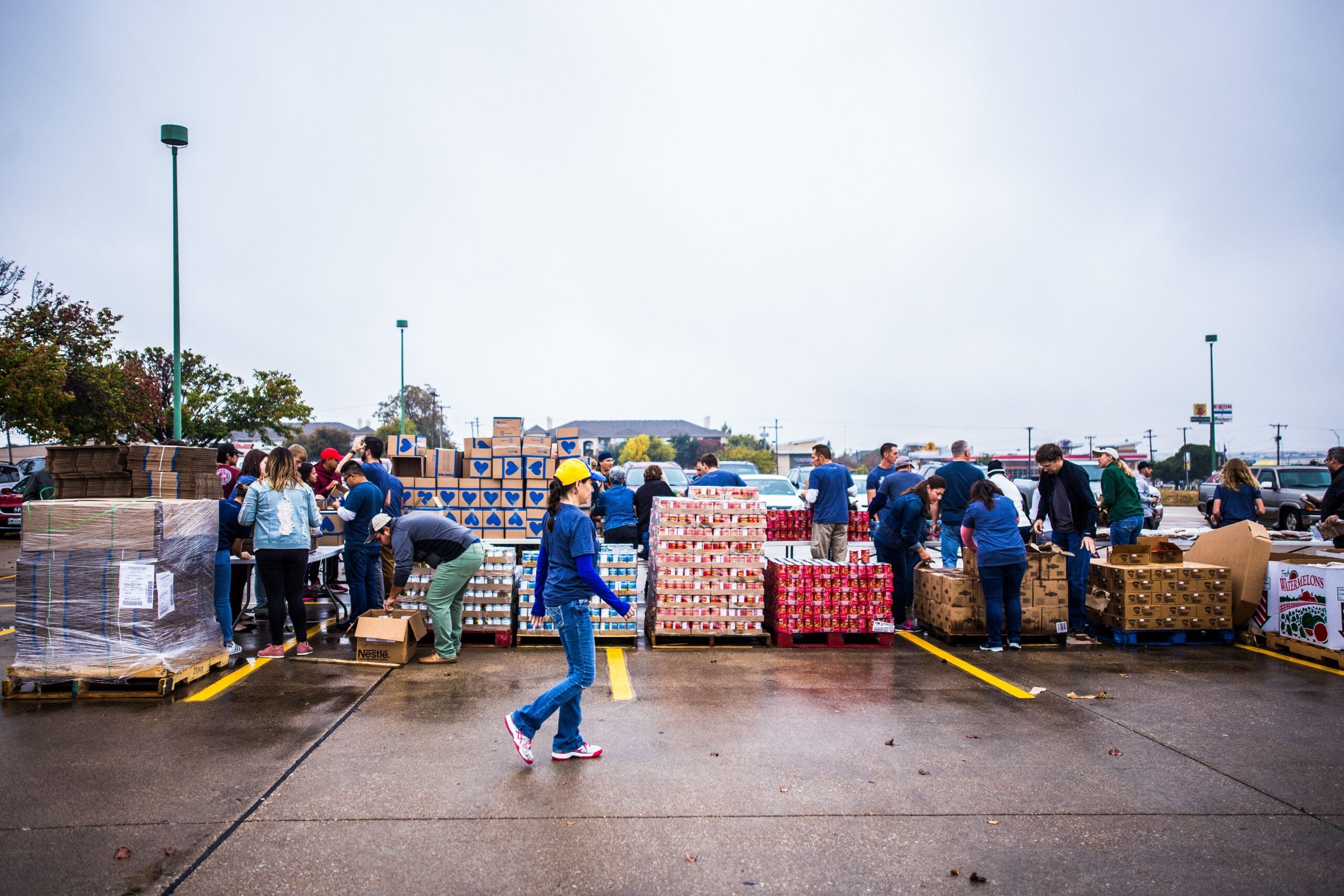 A group of people work together outdoors, sorting and distributing boxes of food on pallets in a parking lot on a cloudy day. One person in a blue shirt and yellow hat walks across the foreground.