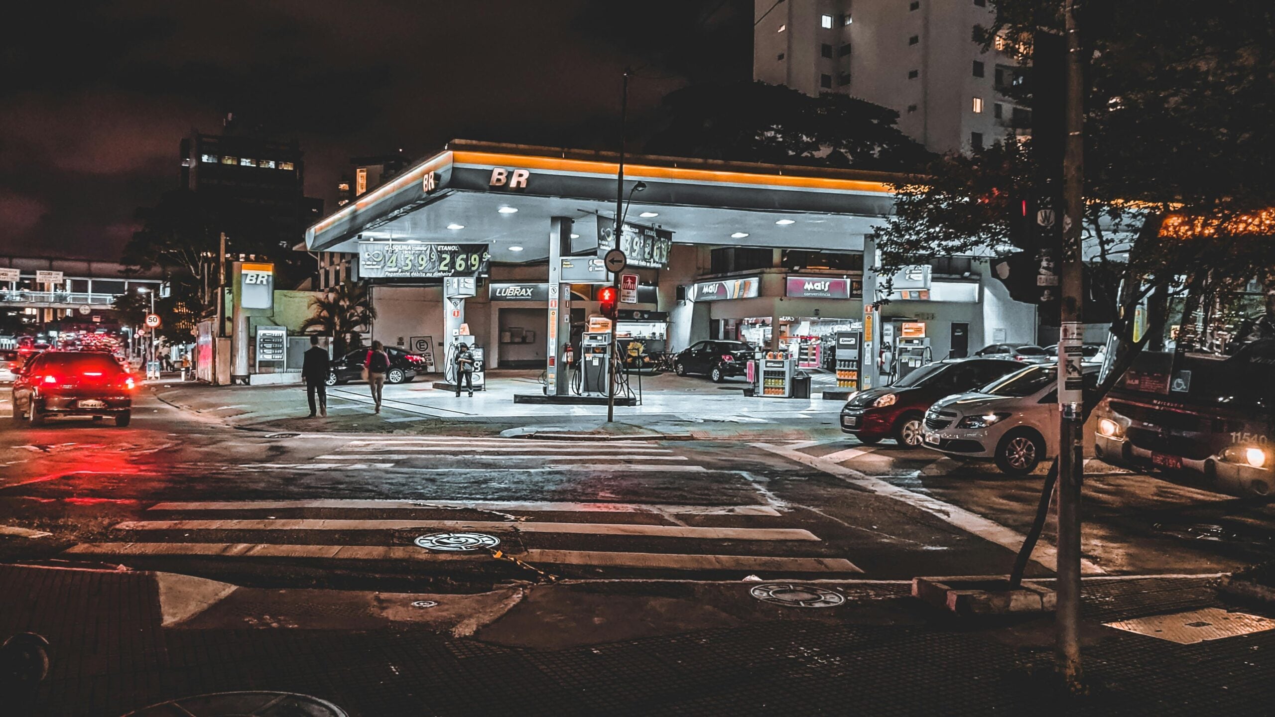 A city gas station at night is brightly lit, with several cars parked at the pumps. The wet street reflects neon signs and headlights. Tall buildings and trees surround the station, and a few people are visible near the entrance.