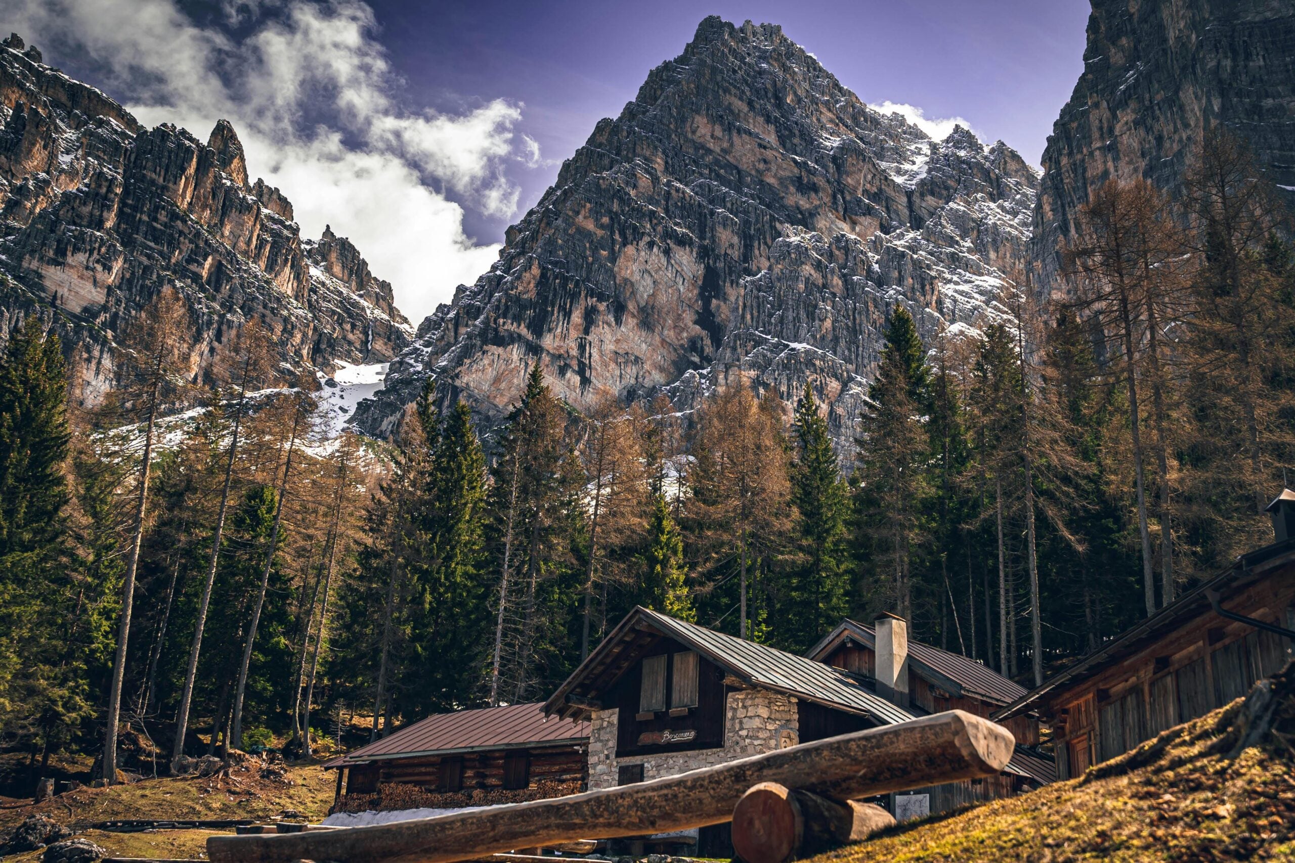 Mountain cabins sit nestled among tall pine trees at the base of steep, rocky, snow-dusted mountains under a partly cloudy sky. A wooden bench lies in the foreground near the rustic buildings.