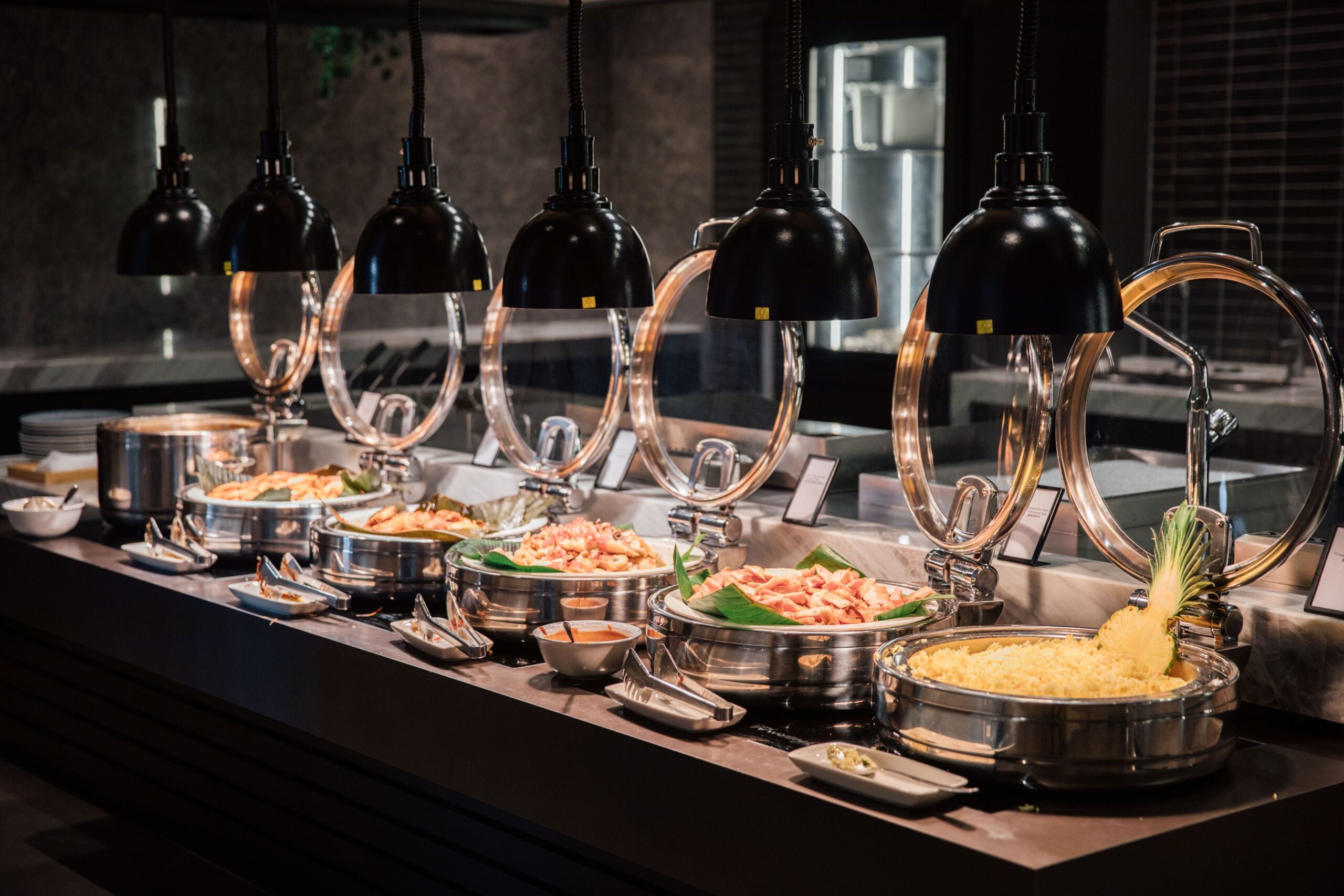 A buffet table displays various hot dishes in metal chafing trays under heat lamps, including pasta, vegetables, and garnished entrees, with serving utensils and small bowls of sauces.