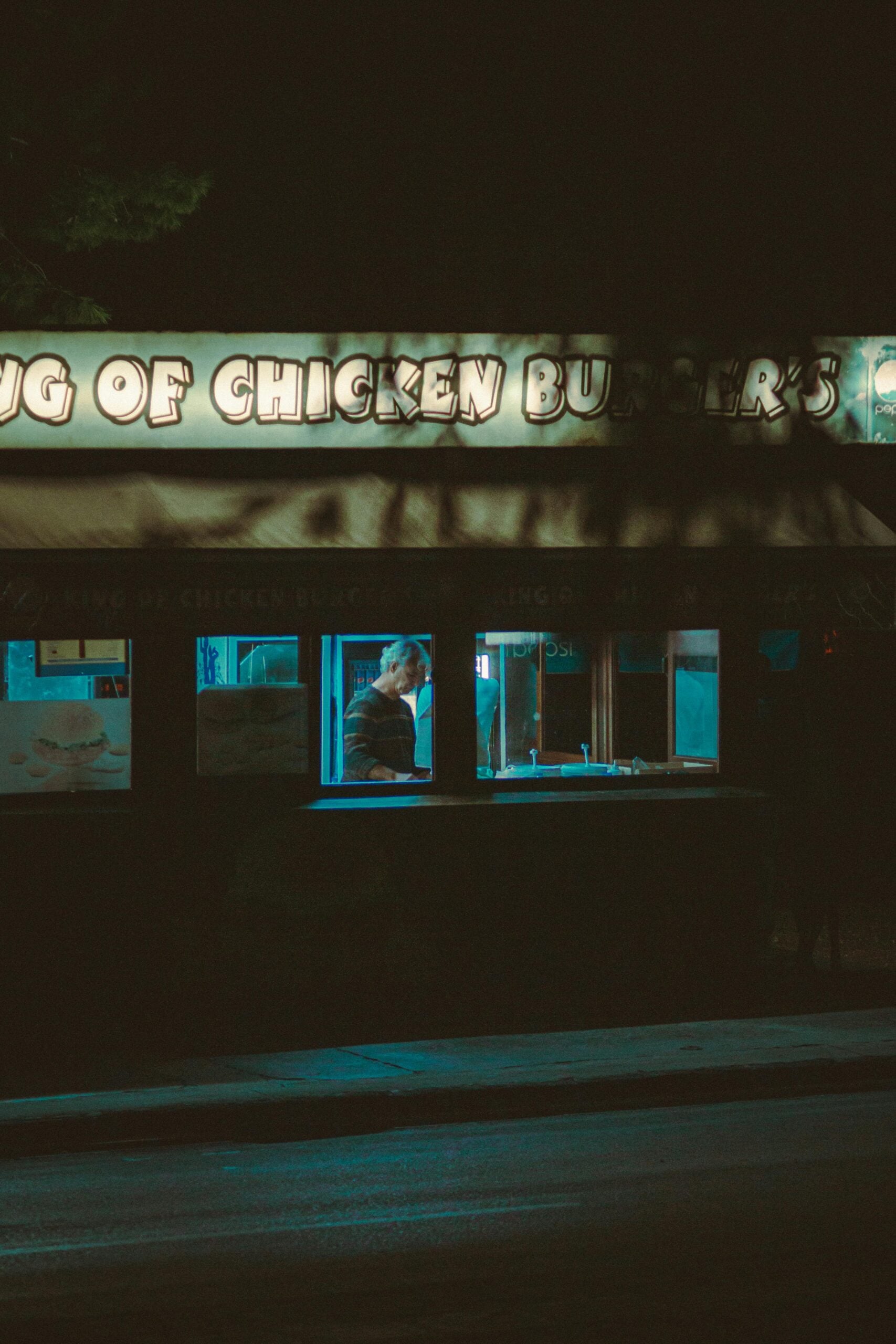 A person stands inside a dimly lit fast food restaurant at night, partially visible through the window under a glowing sign that reads "CHICKEN BURGERS." The street outside is dark and empty.