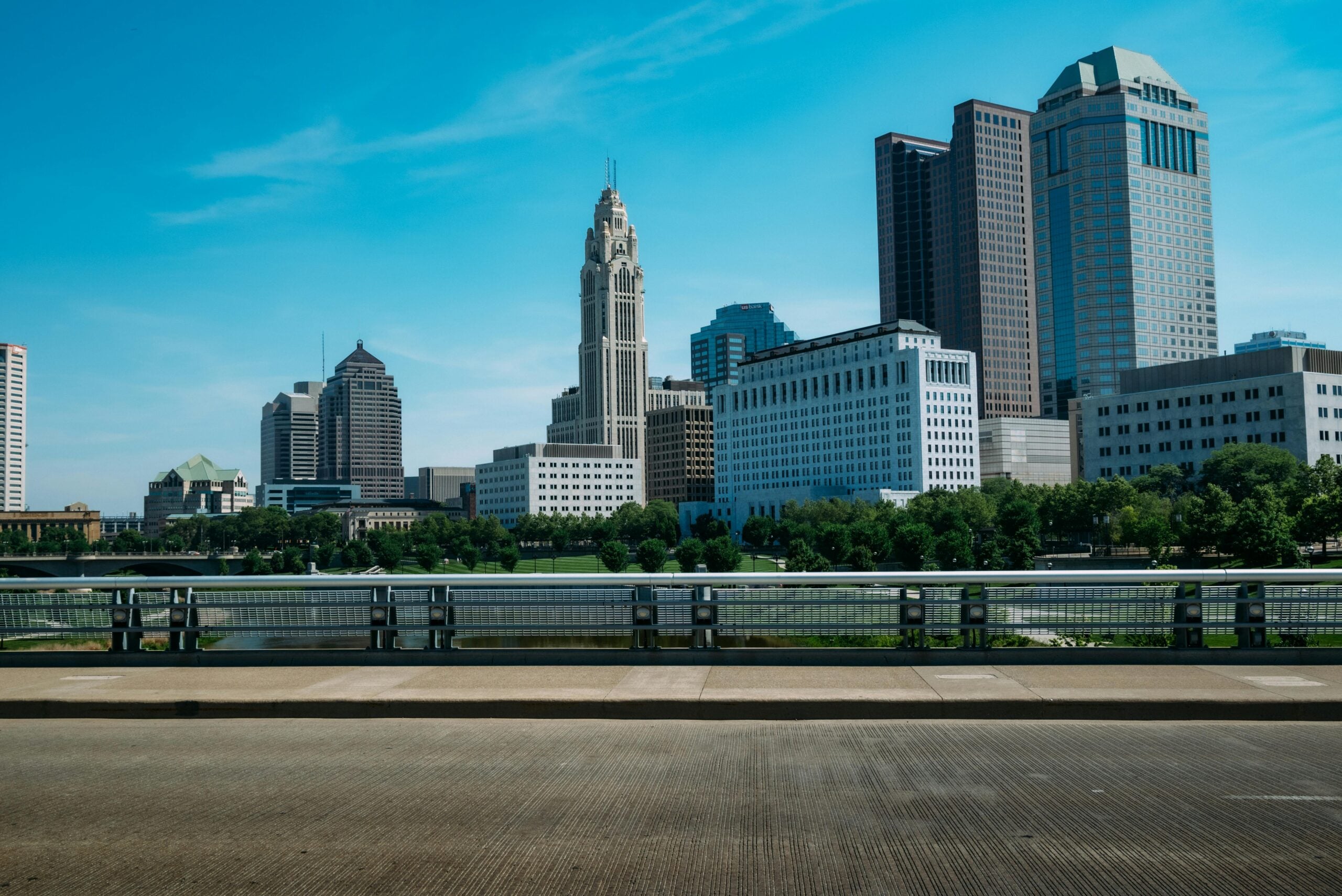 Downtown cityscape with several tall office buildings and skyscrapers under a clear blue sky, seen from across a bridge with metal railings and a road in the foreground. Trees line the area near the buildings.