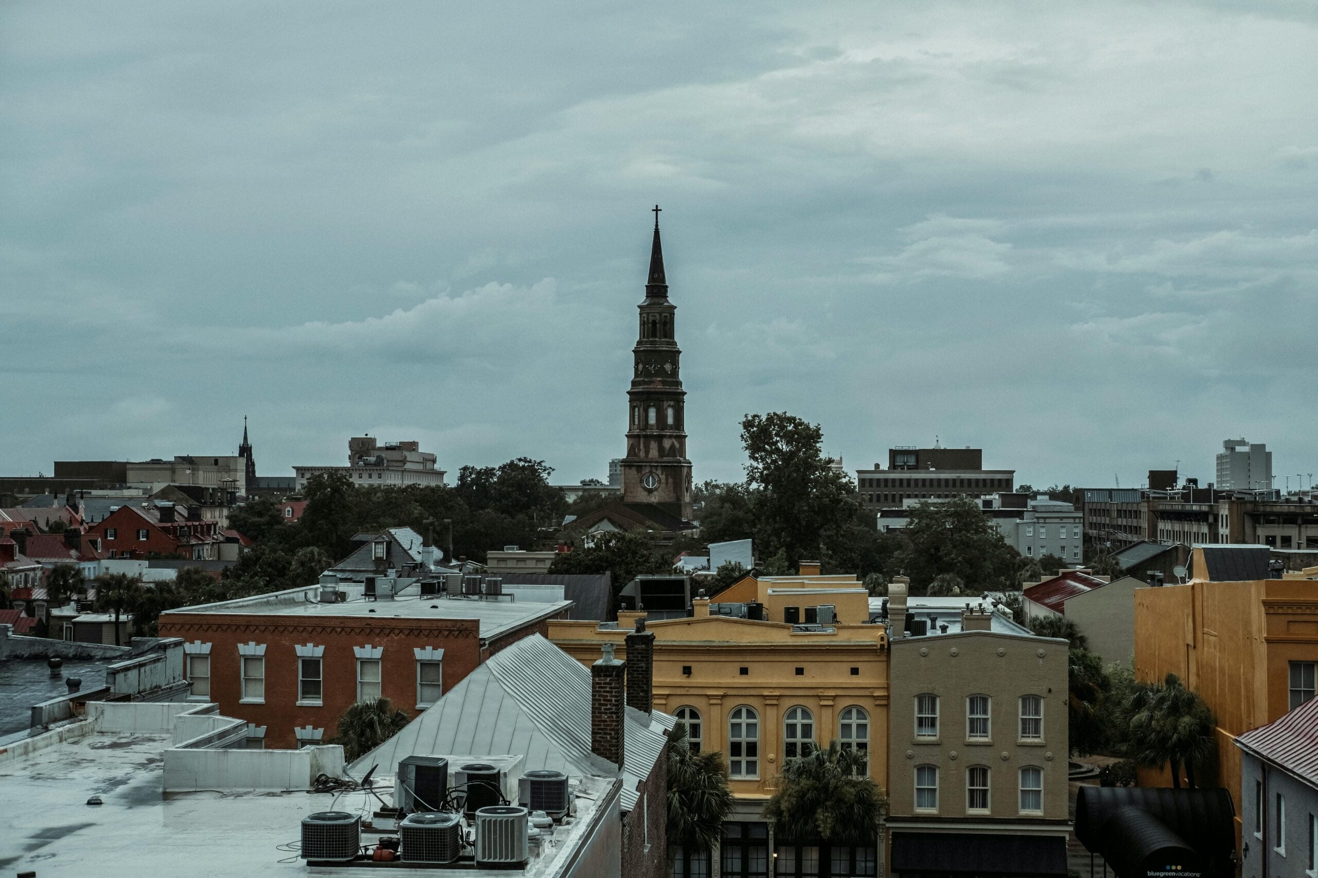 Overcast view of a cityscape with colorful historic buildings in the foreground and a tall church steeple rising among trees and urban structures in the background.