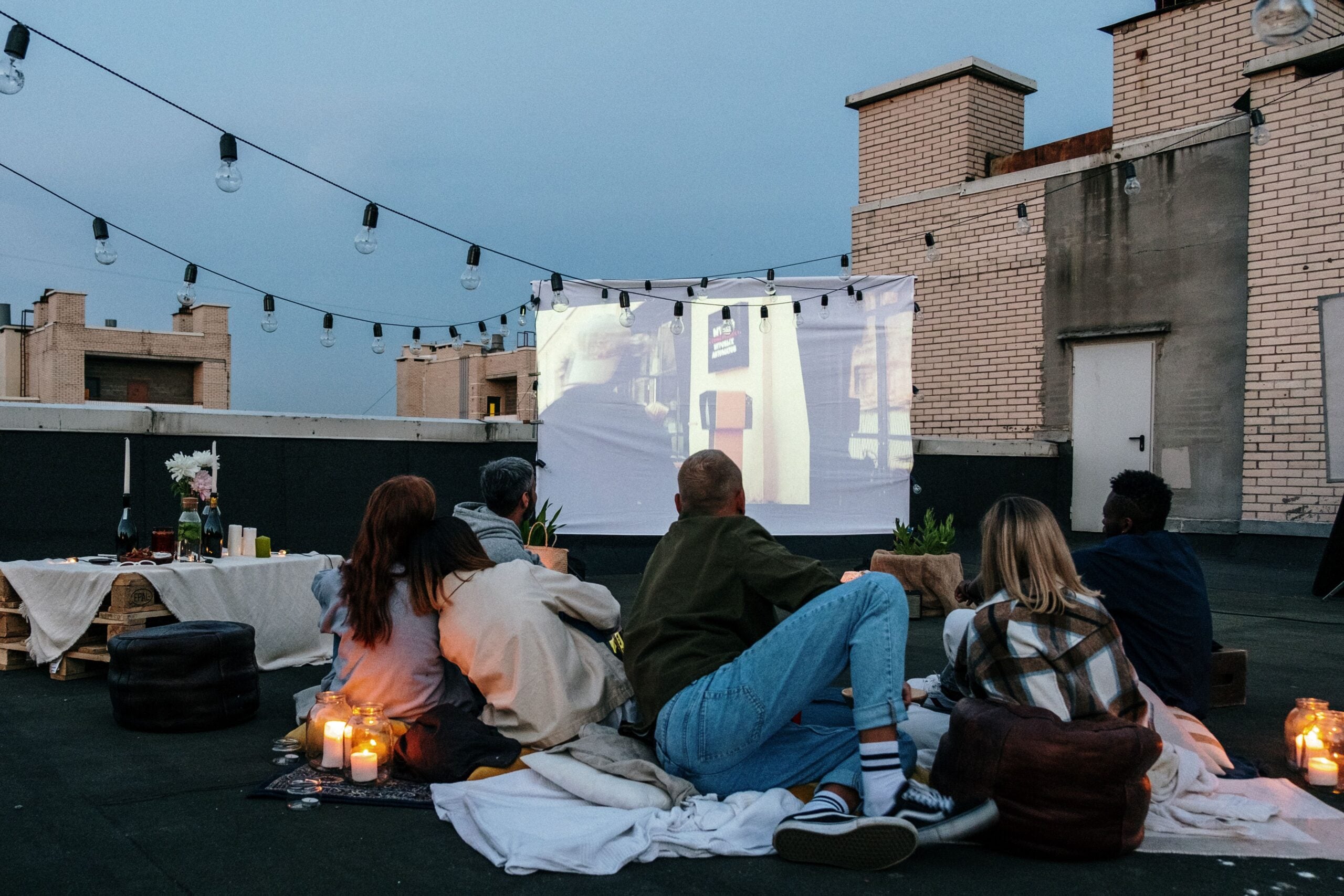 A group of people sit on blankets and cushions on a rooftop, watching a movie projected onto a white screen at dusk. String lights are hung above, and candles and snacks are set on low tables nearby.