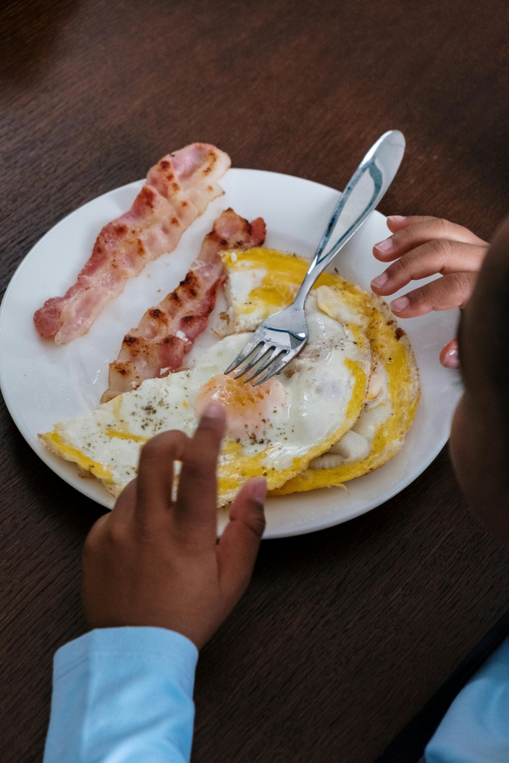 A child’s hands reach for a plate of food with two strips of bacon and two sunny-side-up eggs, with a fork resting on the eggs, on a dark wooden table.