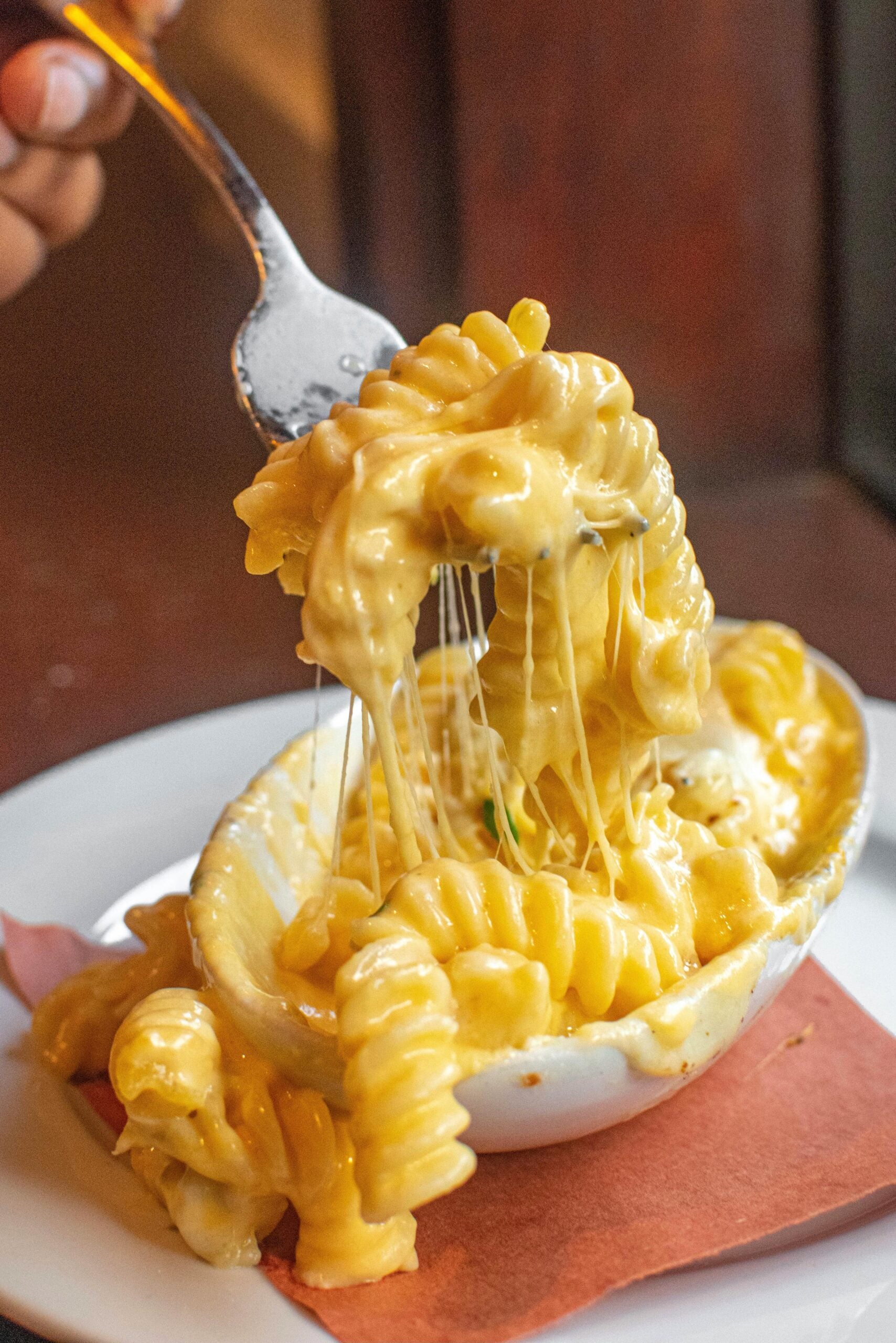 A fork lifts a serving of creamy, cheesy rotini pasta from a white bowl, with melted cheese stretching between the pasta and the bowl, on a white plate with a brown napkin underneath.