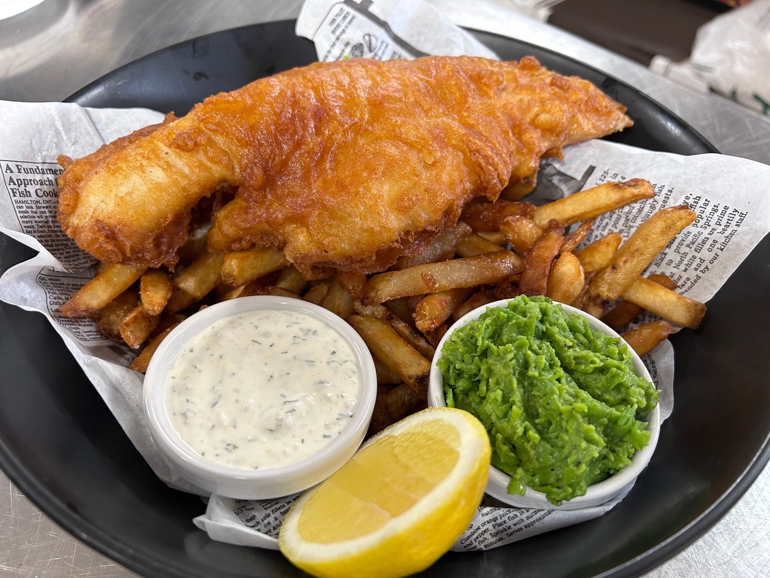 A plate of fish and chips, served on newspaper-style paper with a piece of battered fried fish, golden fries, a wedge of lemon, a bowl of tartar sauce, and a bowl of mushy peas.