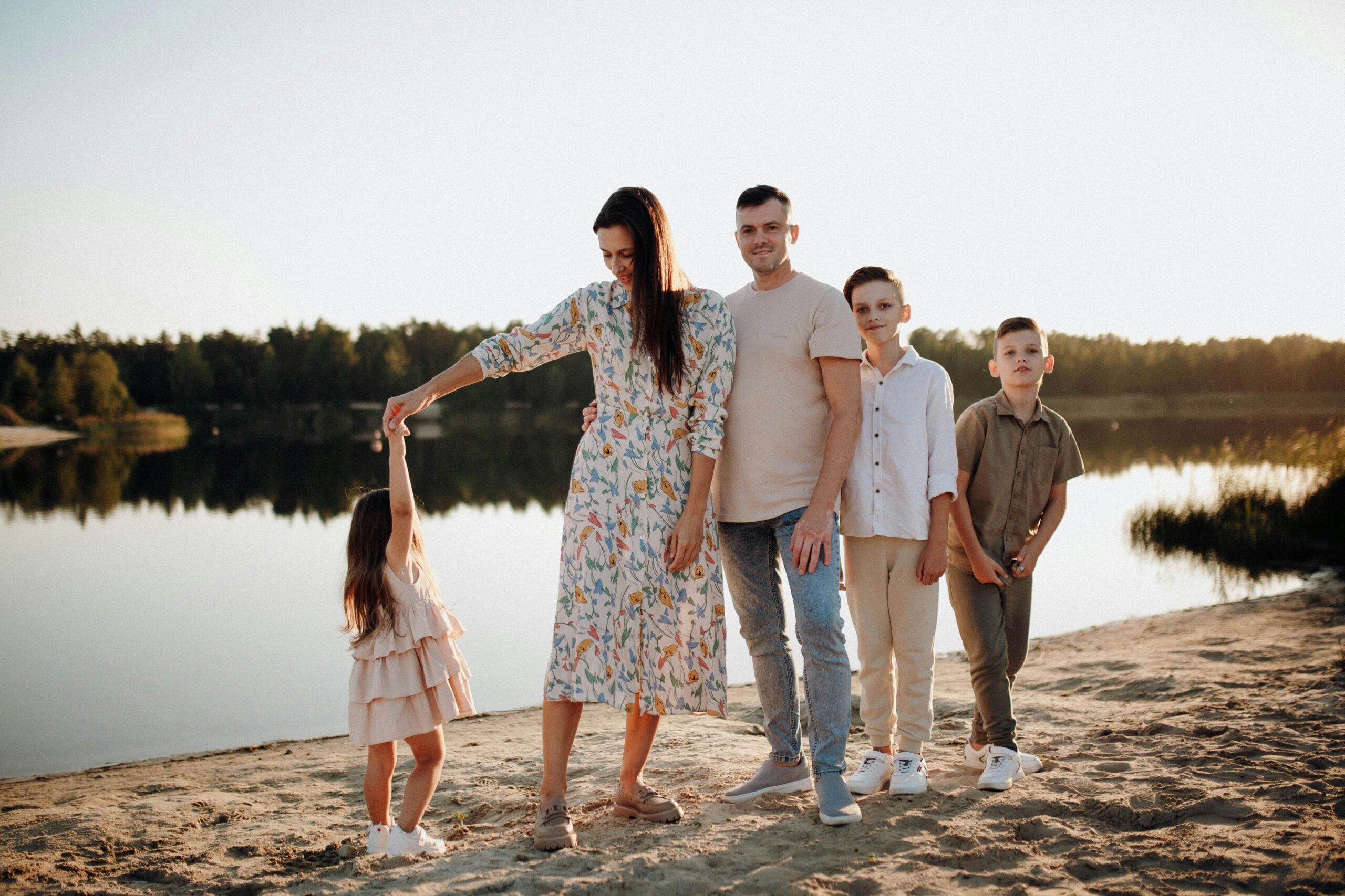 A family of five stands on a sandy shore by a lake at sunset. The mother twirls a young girl, while the father and two boys stand beside them. Trees and water are visible in the background.