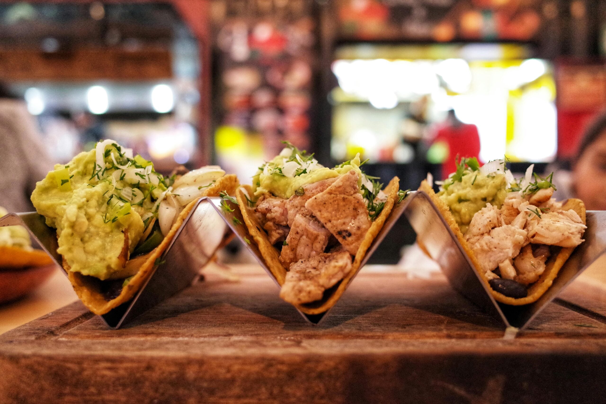 Three tacos filled with meat, guacamole, shredded cheese, and fresh herbs are displayed in a metal taco holder on a wooden surface, with a blurred restaurant background.
