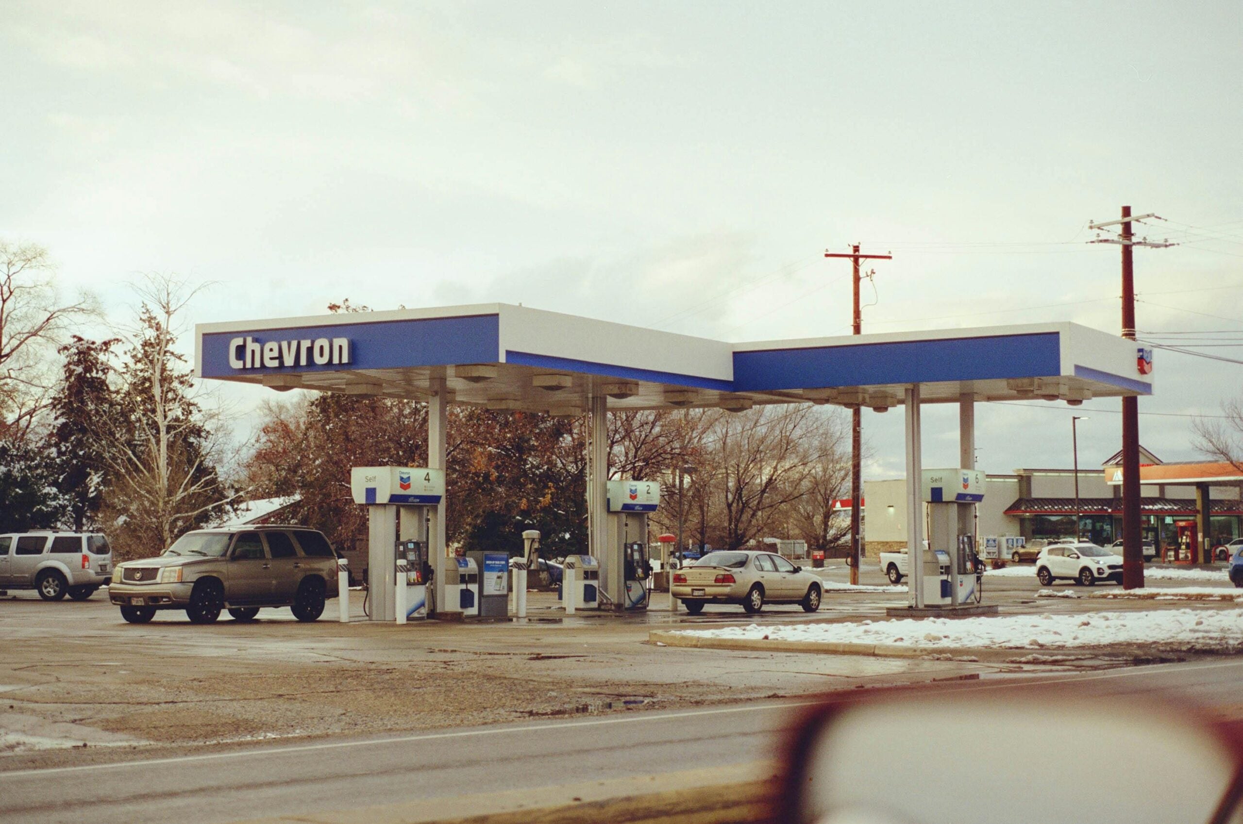 A Chevron gas station with several cars parked at the pumps on a cloudy day. There is some snow on the ground and trees in the background, with other vehicles and buildings visible nearby.