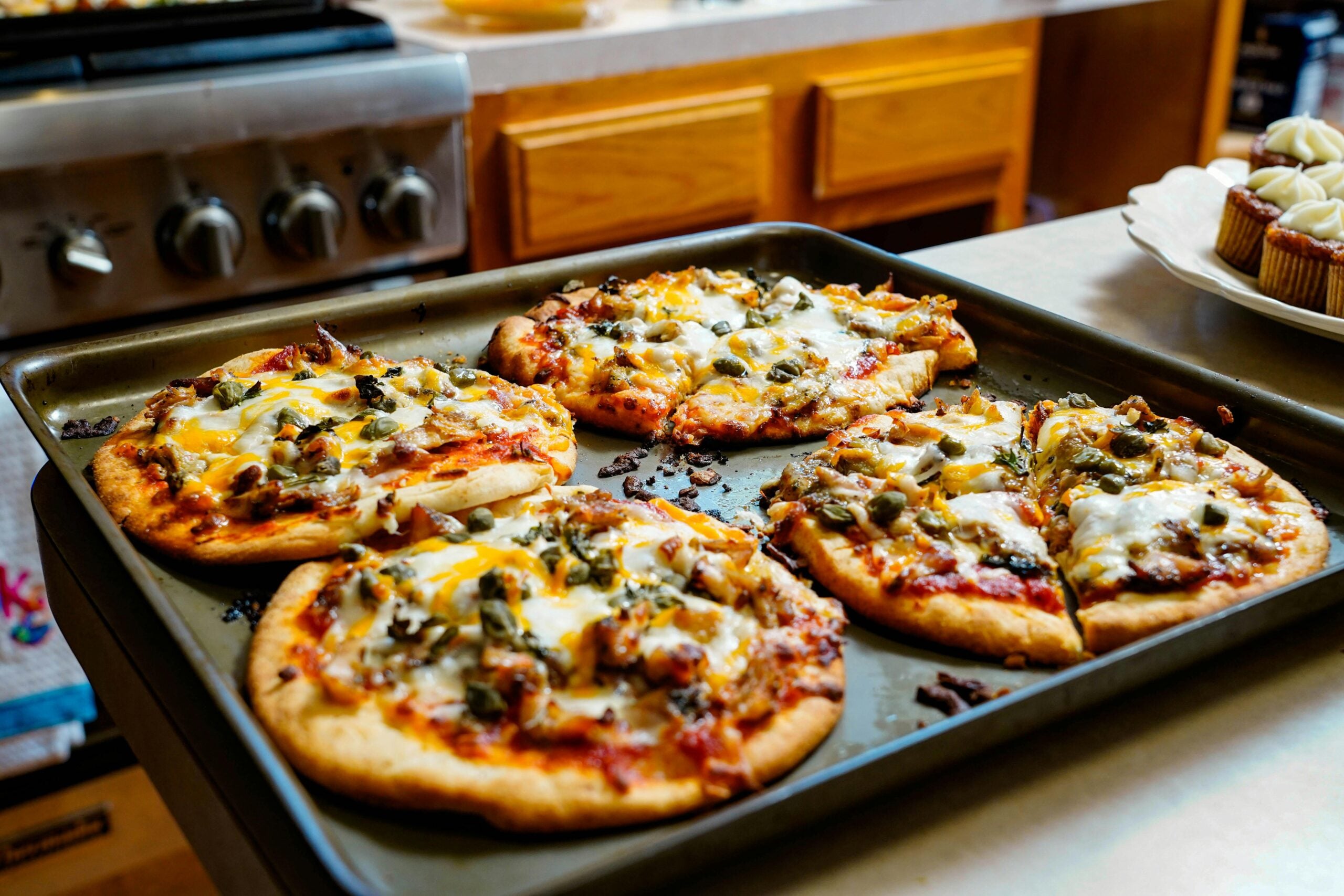 A baking sheet with five small homemade pizzas topped with melted cheese and vegetables sits on a kitchen counter, with cupcakes and cooking utensils visible in the background.