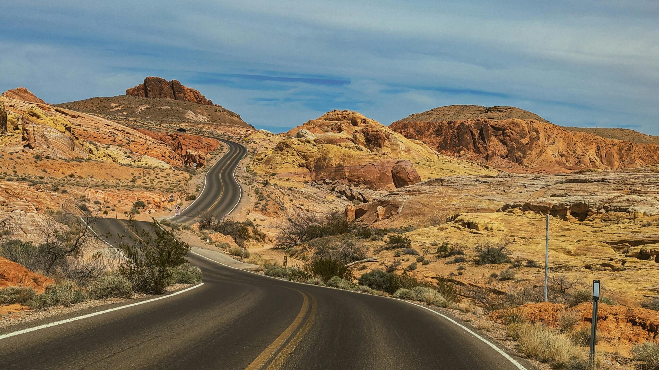 A winding road stretches through a desert landscape with colorful, rocky hills and sparse vegetation under a blue sky with wispy clouds.