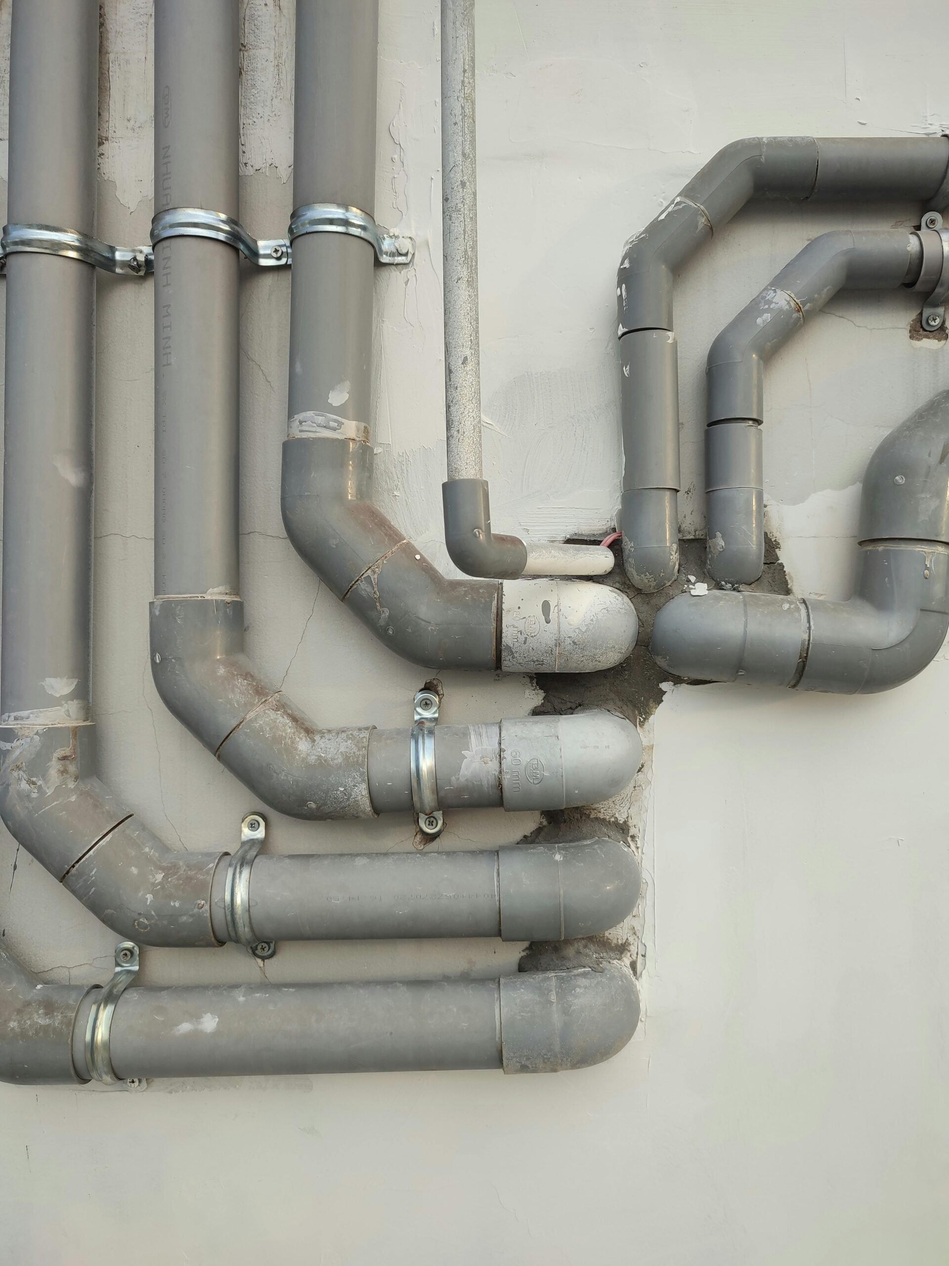 Gray pipes with metal clamps are attached to a pale wall, running horizontally and vertically with several bends and joints, creating a geometric pattern. Some pipes have scratches and patches.