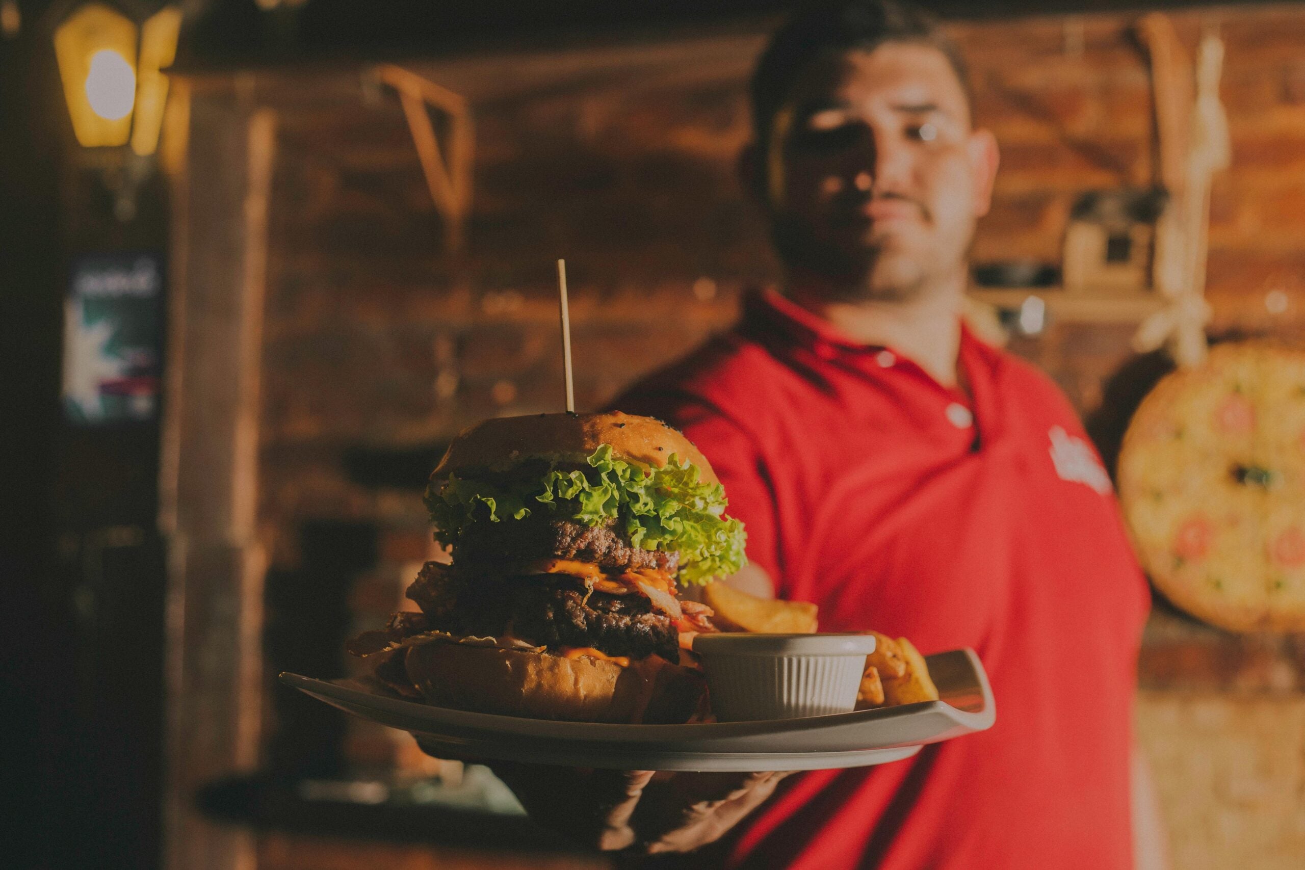 A man in a red polo shirt presents a plate with a large burger, fries, and a small cup of sauce. The background is dimly lit with wooden decor and a pizza on the wall.