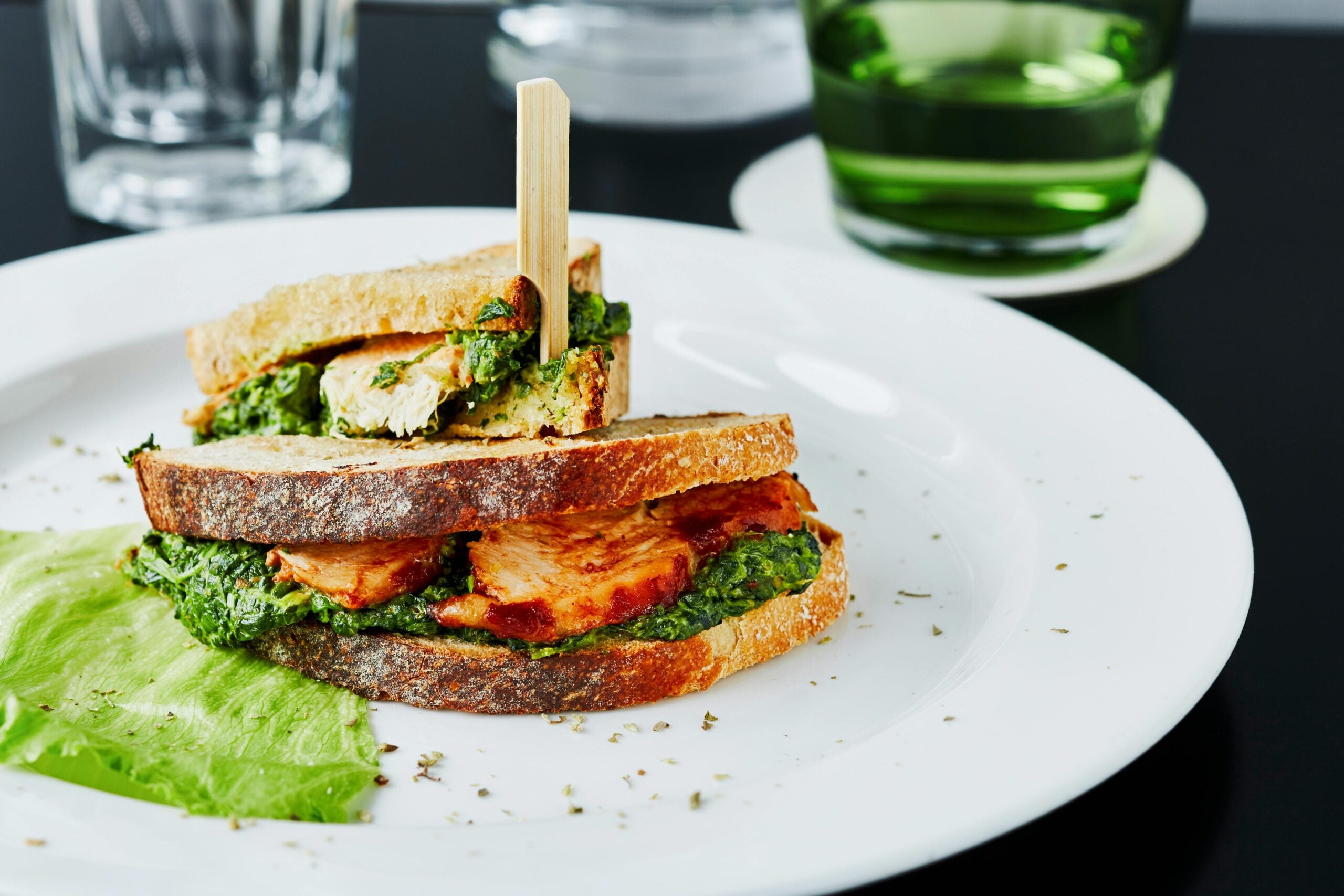 A sandwich with grilled chicken, green pesto, and leafy greens is served on rustic bread, secured with a wooden pick on a white plate, garnished with a lettuce leaf and herbs. Glasses and a green cup are in the background.