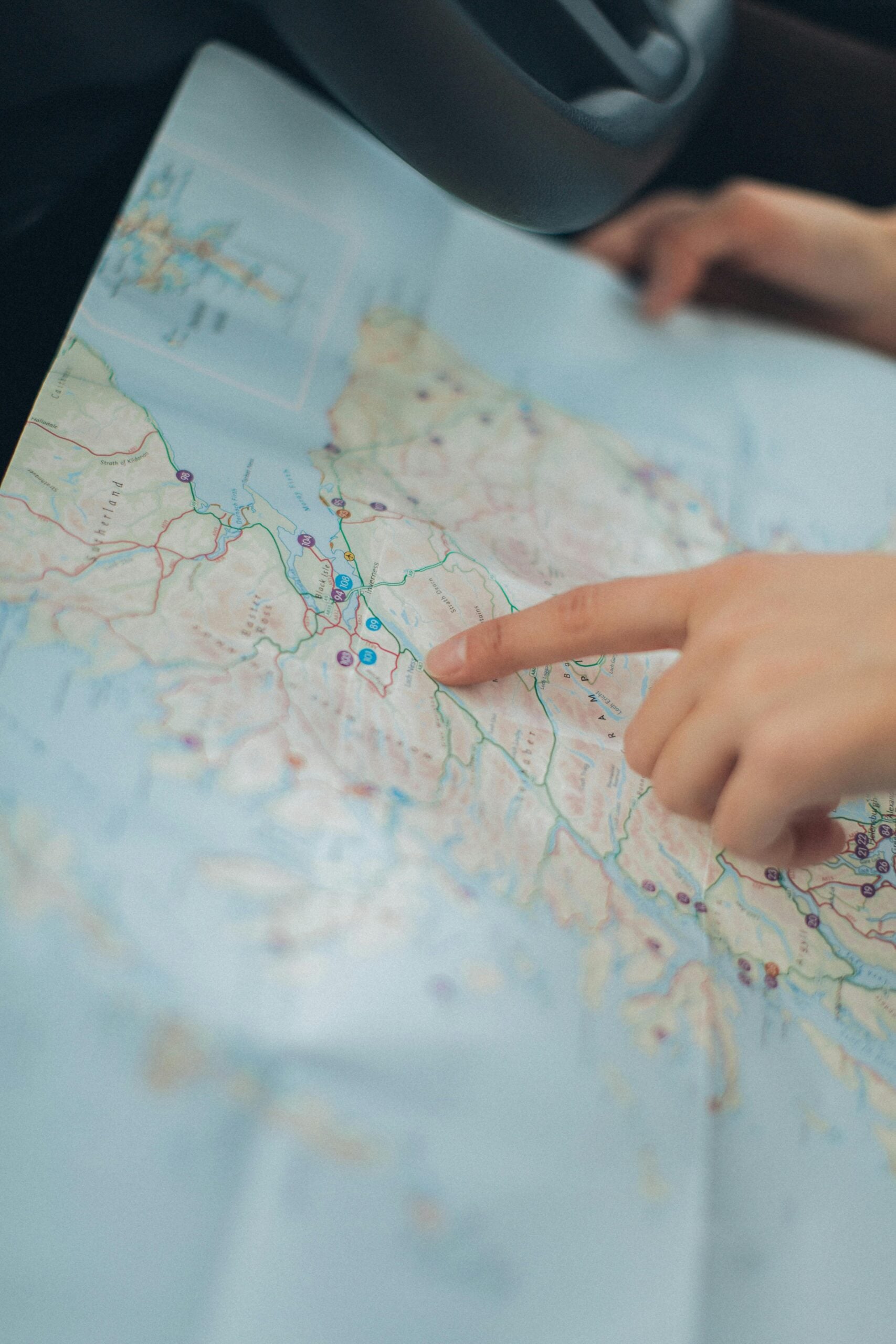 A person points to a location on a paper map laid out inside a car, with the steering wheel and another hand visible nearby.