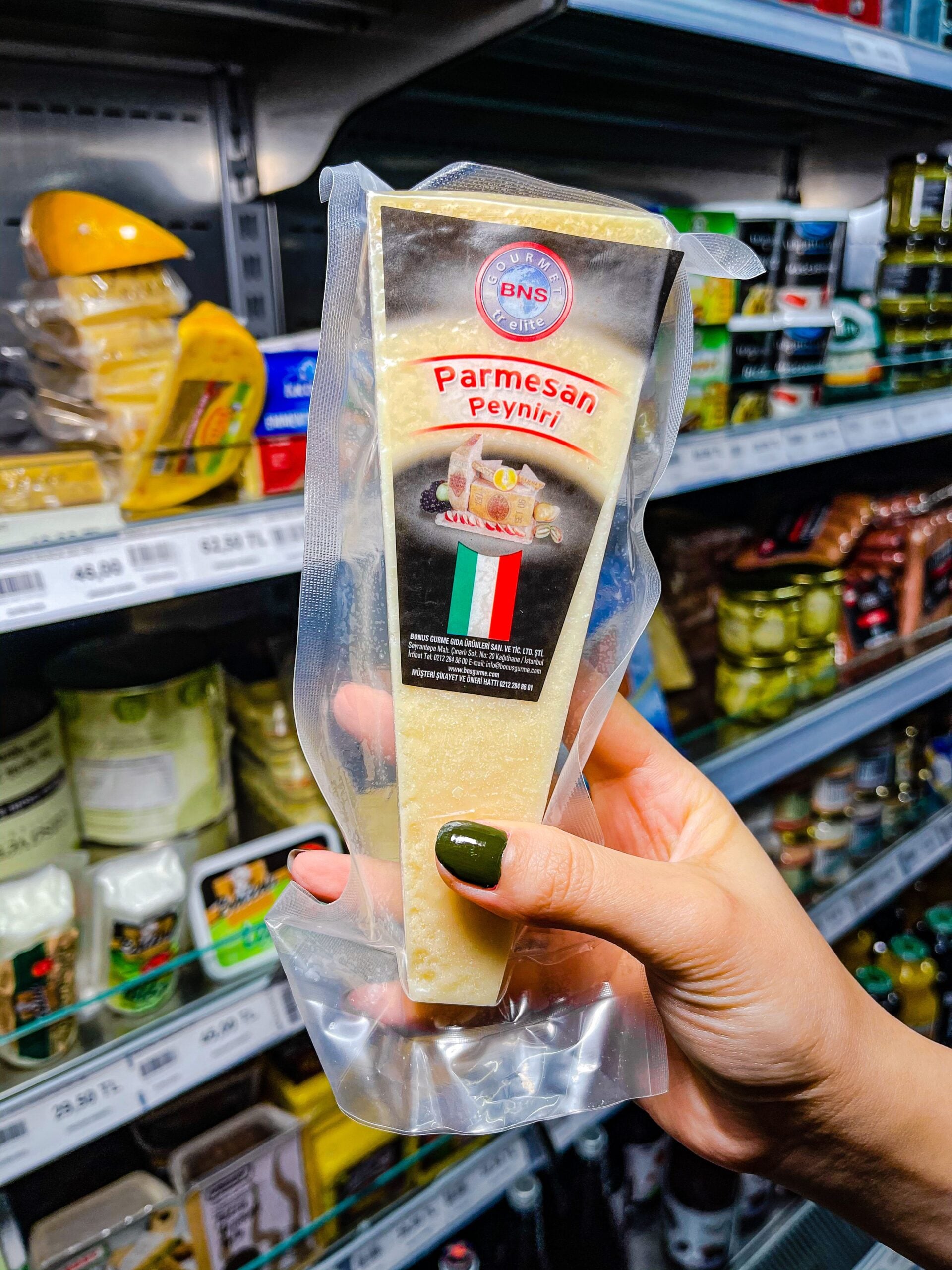 A hand with dark green nail polish holds a wedge of packaged Parmesan cheese in a grocery store, with shelves of various cheeses and jars in the background.