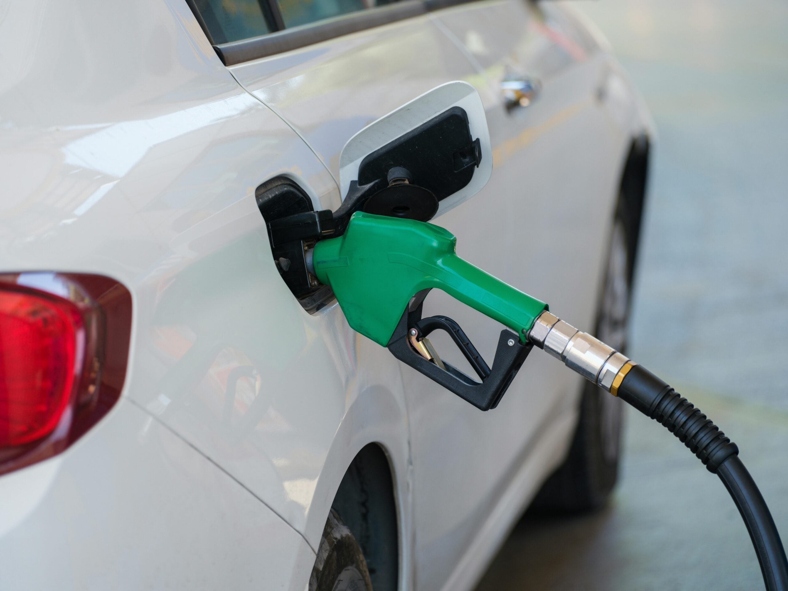 A green fuel nozzle is inserted into the gas tank of a white car at a gas station. The car’s fuel door is open, and the nozzle is active, indicating the car is being refueled.