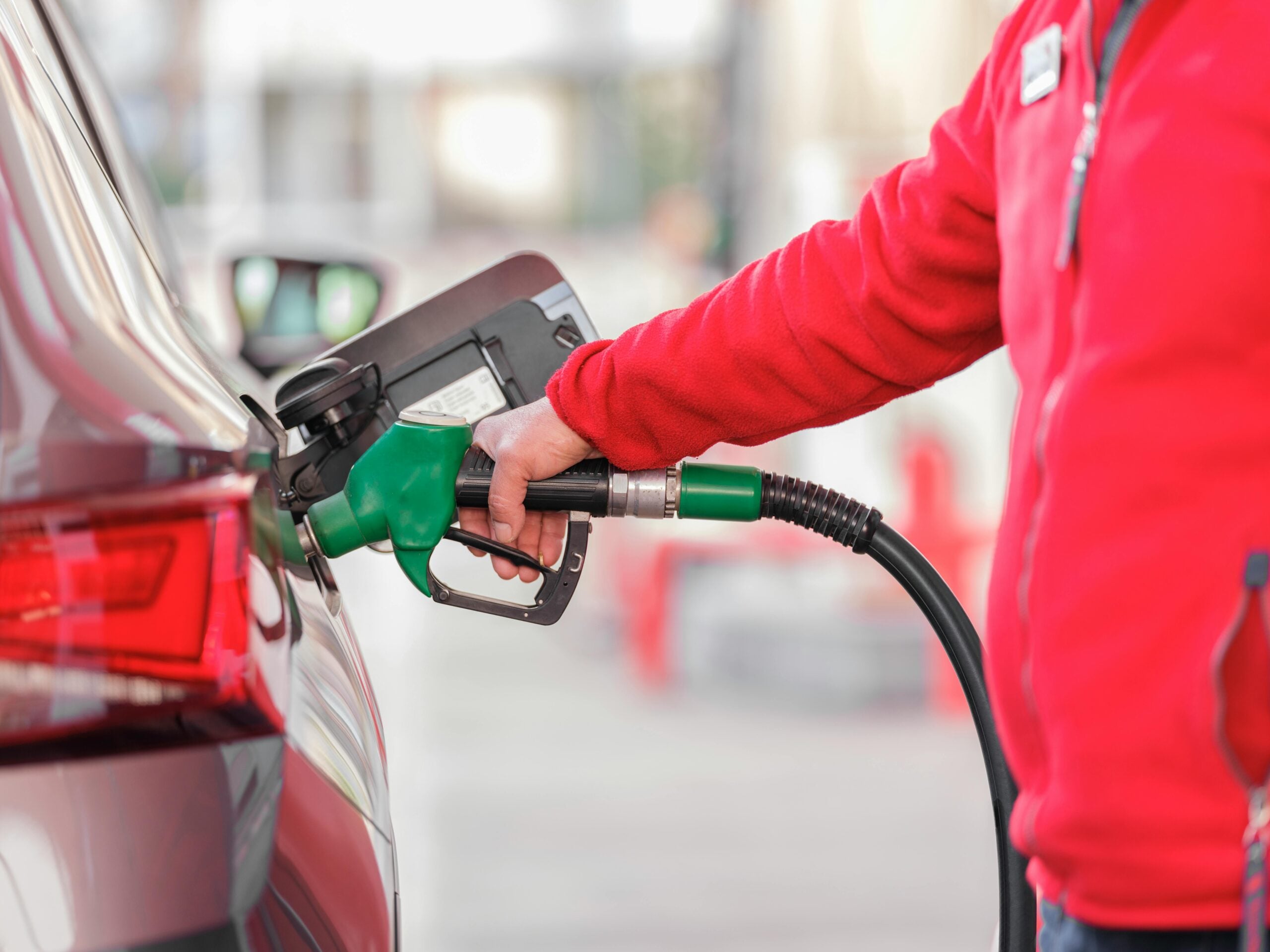 A person in a red jacket is refueling a car at a gas station, holding a green fuel nozzle inserted into the vehicle’s tank. The background is blurred.