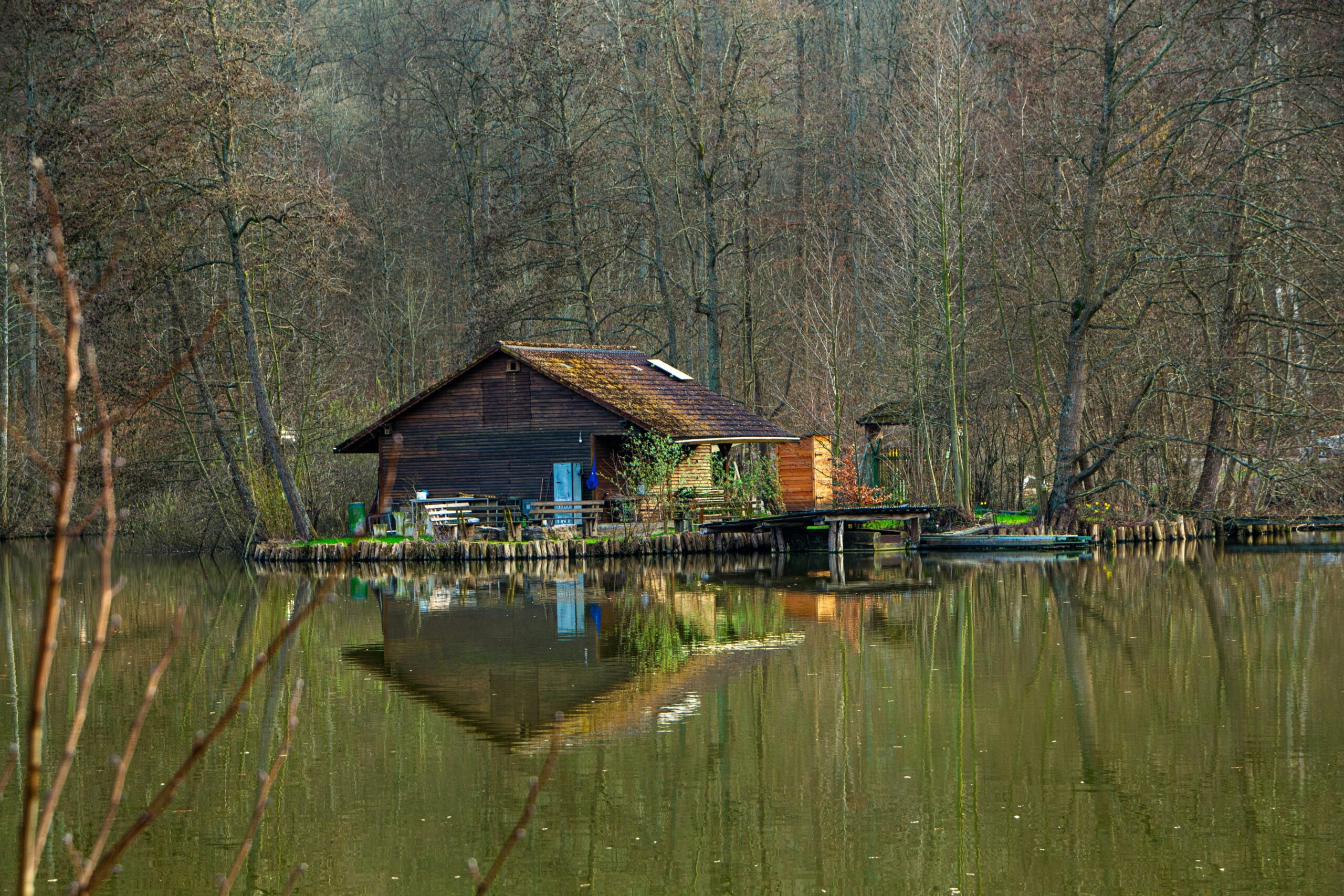 A small wooden cabin sits on the edge of a calm lake, surrounded by bare trees. The cabin and trees are reflected clearly in the still water, creating a peaceful, secluded scene in nature.
