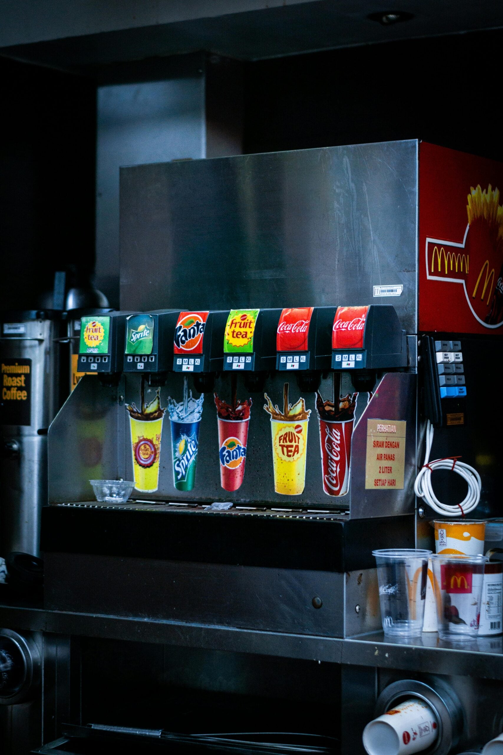 A McDonald’s self-serve soda fountain machine dispensing drinks, with options for Sprite, Fanta, Fruit Tea, and Coca-Cola. Several cups are placed below the dispensers, and empty cups are stacked on the side.