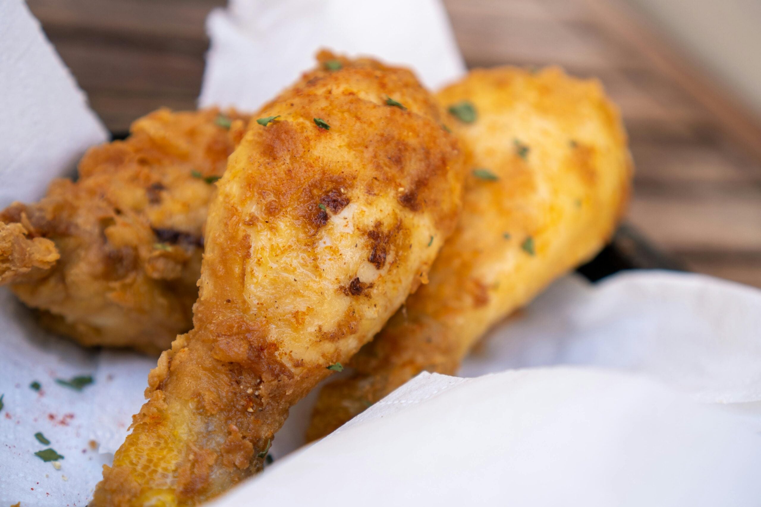 Close-up of crispy fried chicken drumsticks resting on white paper towels, garnished with small bits of green herbs. The chicken has a golden-brown, crunchy coating.
