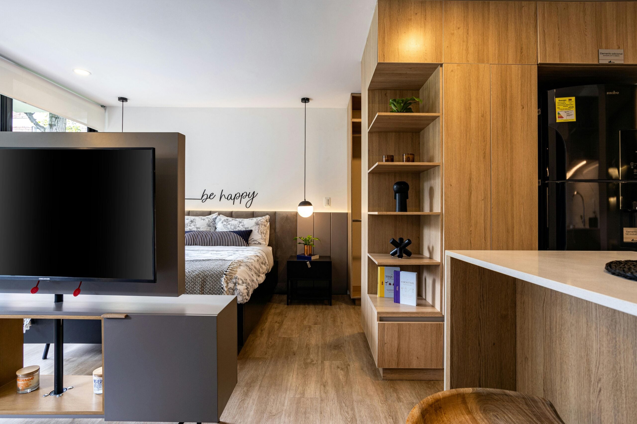 Modern studio apartment with wood accents, a bed next to a wall with "be happy" text, open shelving, a flat-screen TV, and a kitchen area with a counter and refrigerator. Natural light fills the space.
