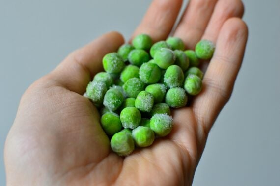 A close-up of a person's hand holding a handful of frozen green peas, with visible ice crystals on the peas. The background is plain and out of focus.