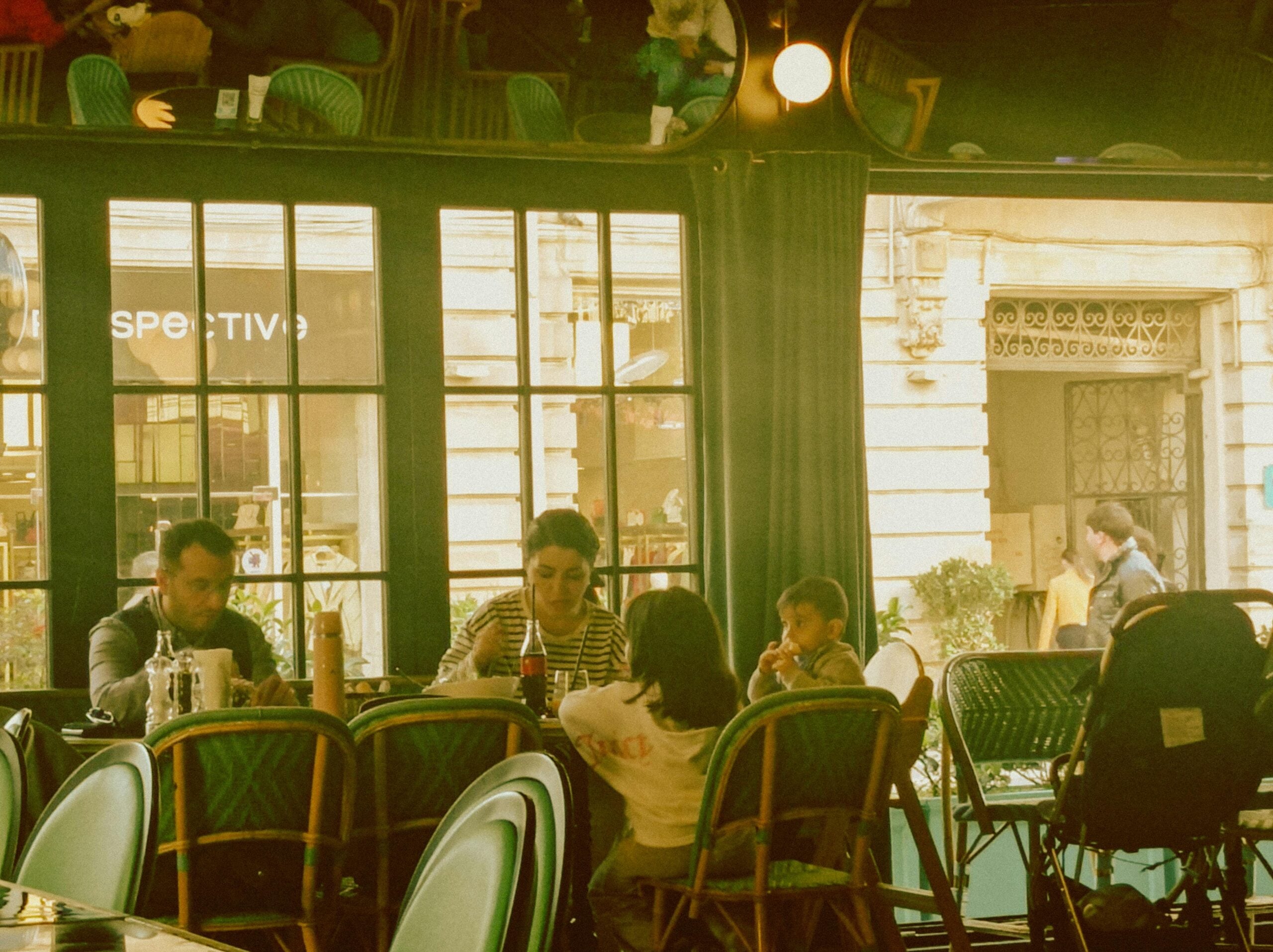 A family of four sits around a table at a cozy café with large windows, enjoying a meal together. The scene is warmly lit, and there are empty chairs and a stroller nearby. A street and pedestrians are visible outside.