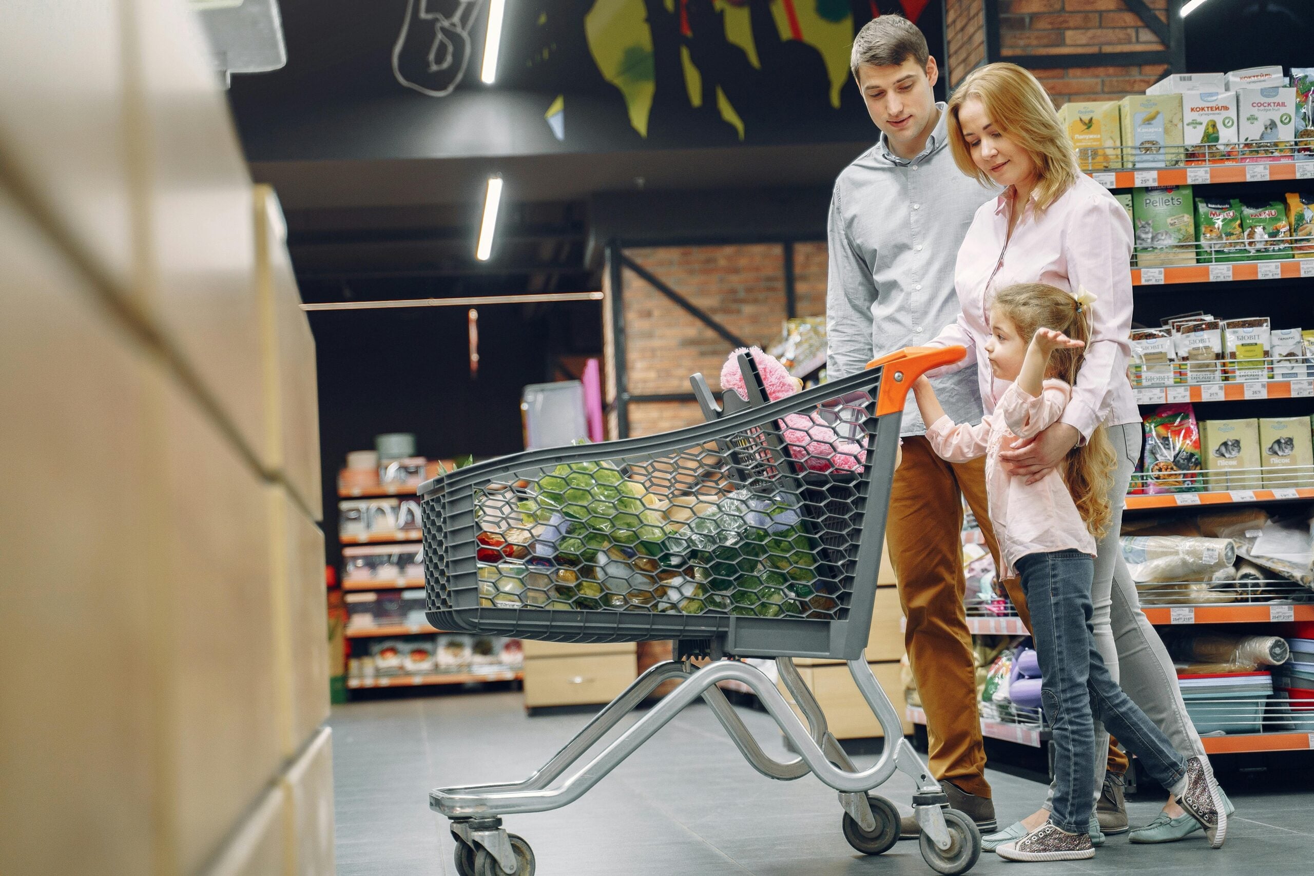 A family with a man, woman, and young girl stands together in a grocery store aisle, holding hands near a shopping cart filled with groceries, surrounded by shelves of products.