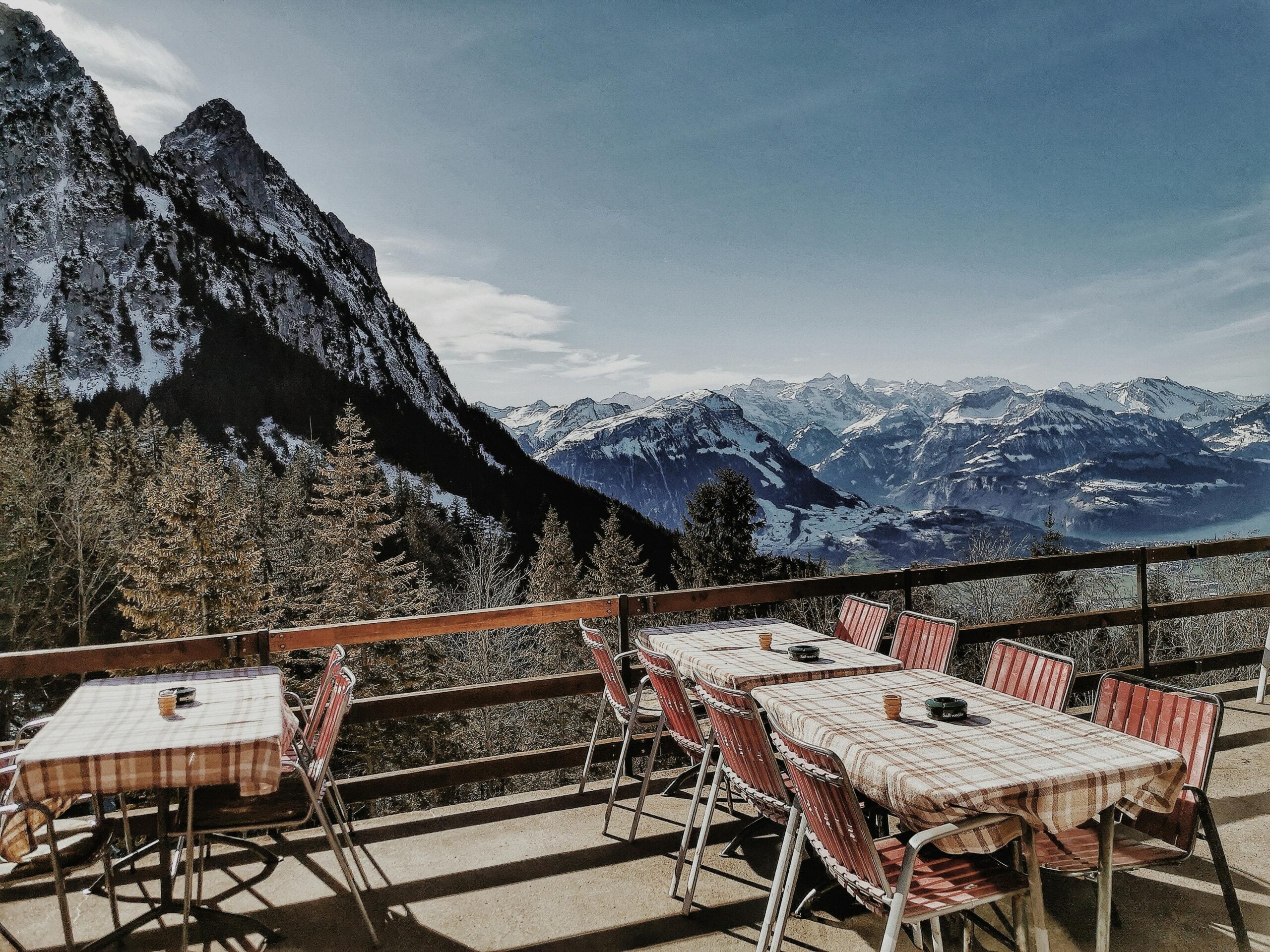 Outdoor terrace with empty tables and chairs, overlooking snow-covered mountains and pine trees under a clear blue sky.