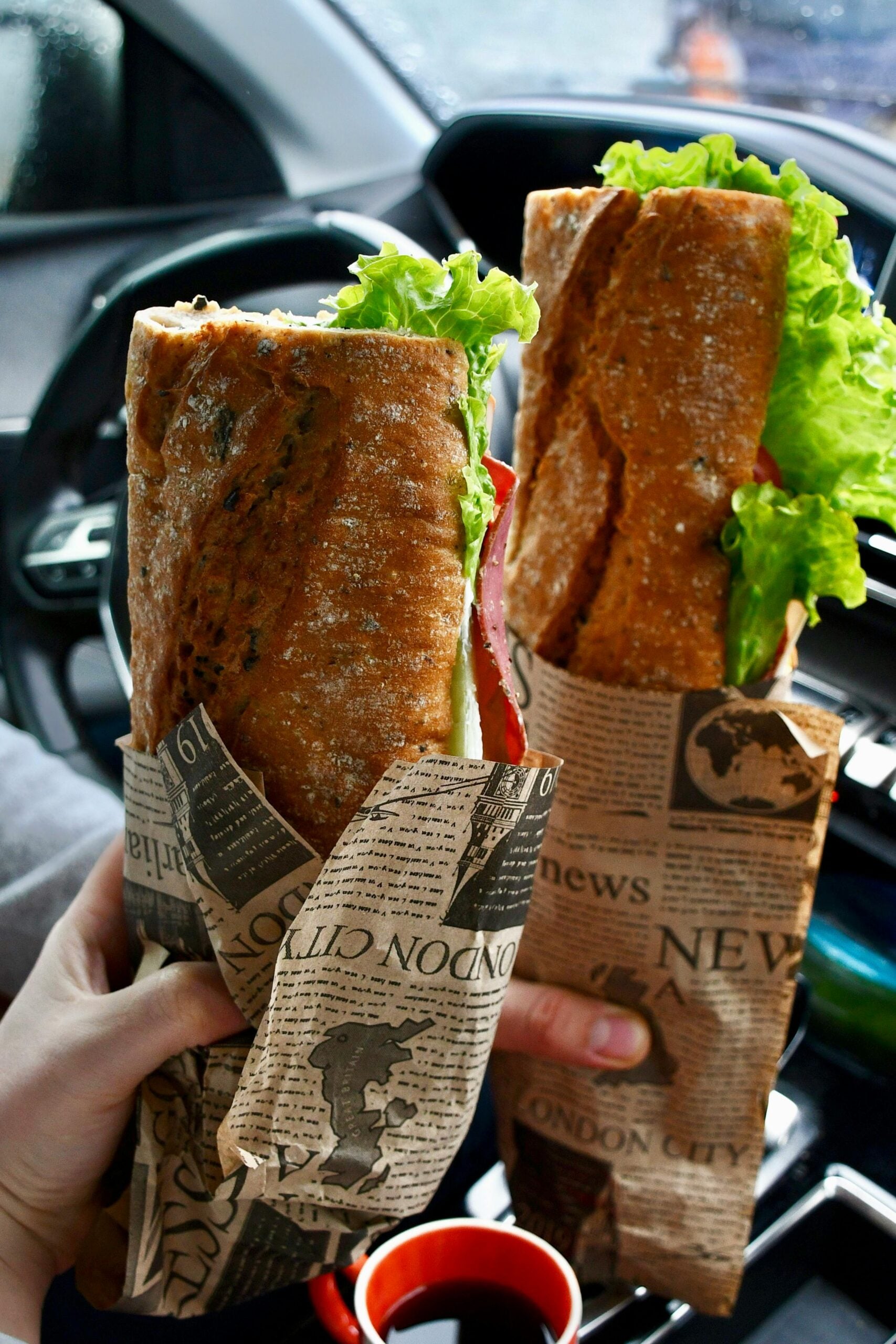Two hands hold large baguette sandwiches wrapped in newspaper print paper, filled with lettuce and other ingredients, inside a car.