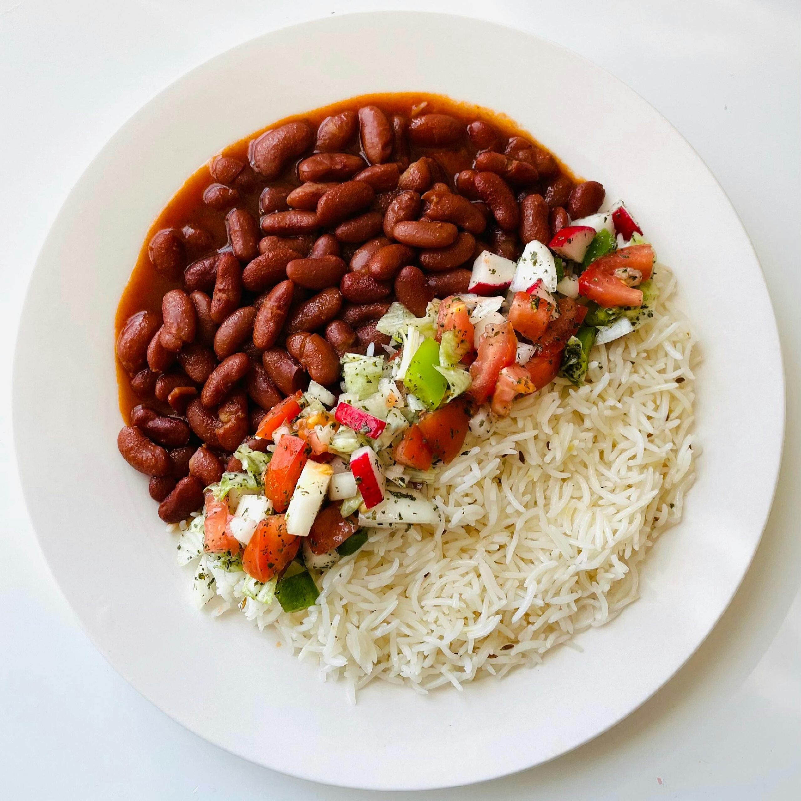 A white plate with cooked white rice, red kidney beans in sauce, and a colorful salad made of diced tomatoes, cucumbers, radishes, and cheese, arranged in separate sections.