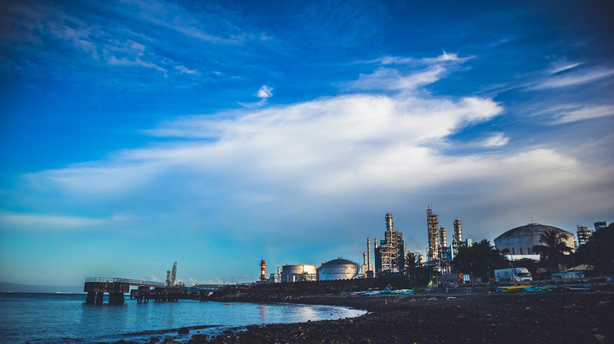Industrial refinery complex with large storage tanks and towers near a shoreline under a vibrant blue sky with scattered clouds; calm water and rocky beach in the foreground.