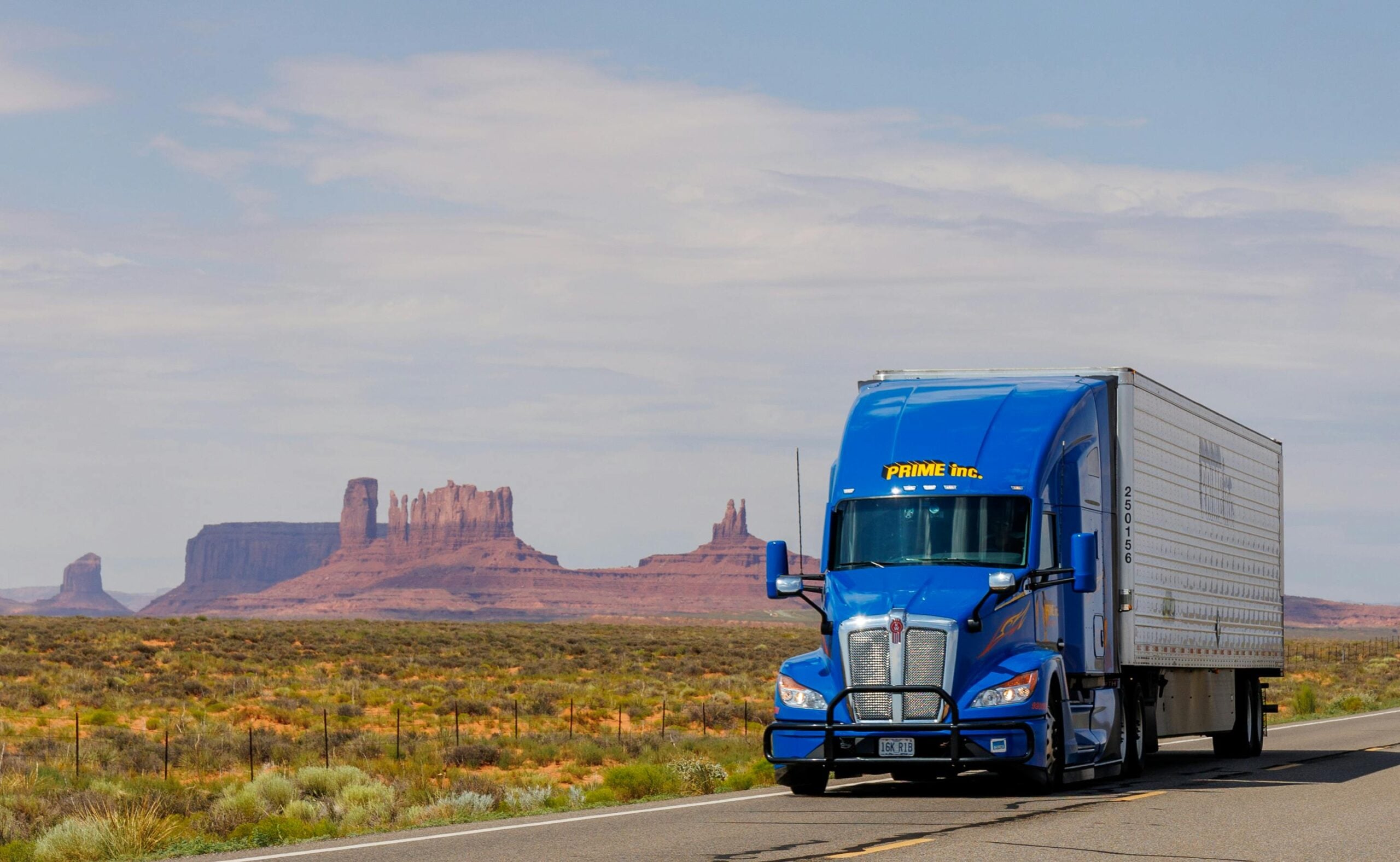 A blue semi-truck with "PRIME INC." on its cab drives on a highway through a desert landscape with mesas and rock formations in the background, under a partly cloudy sky.