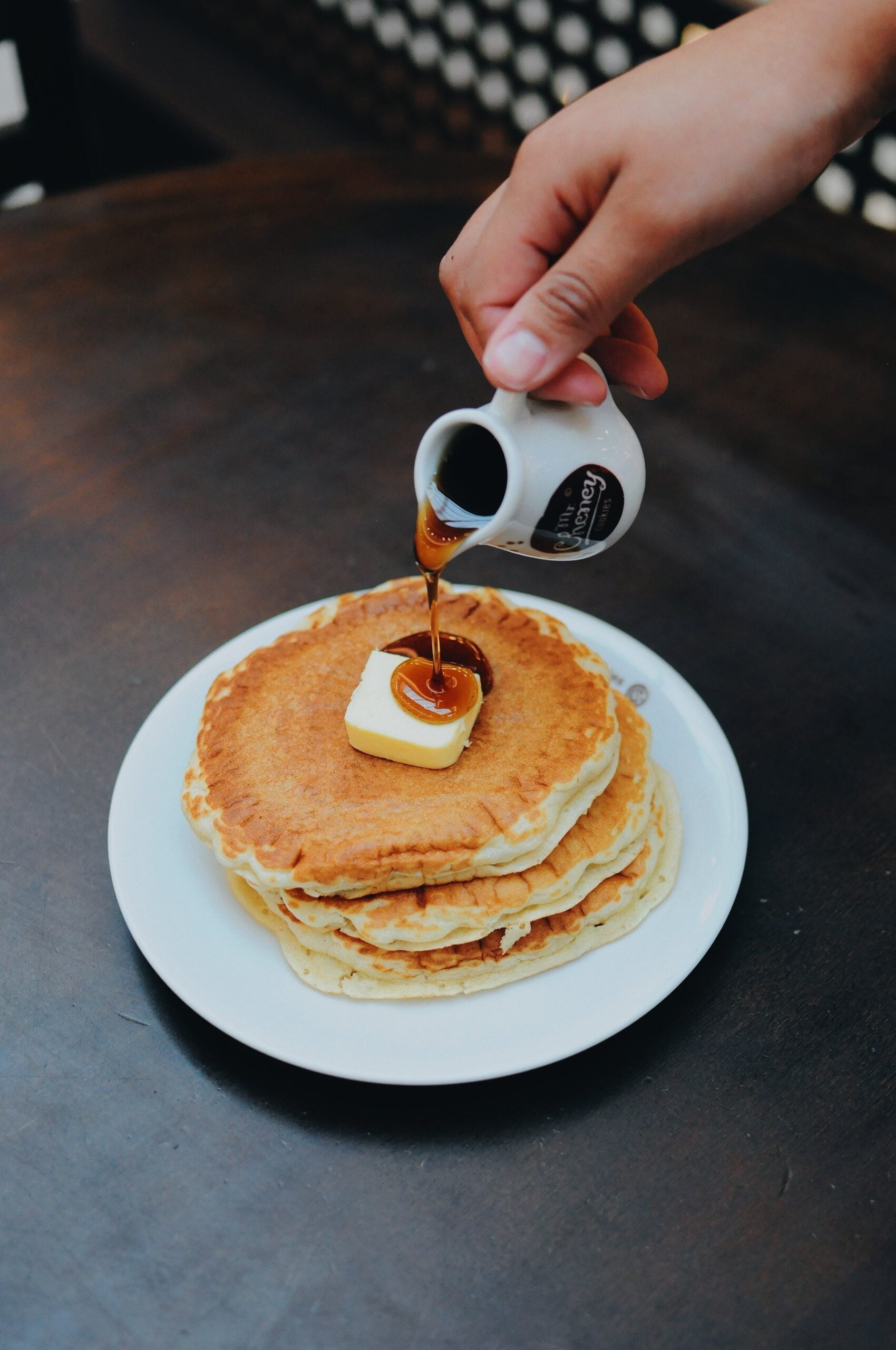 A hand pours syrup from a small pitcher onto a stack of pancakes with a pat of butter on top, served on a white plate against a dark background.