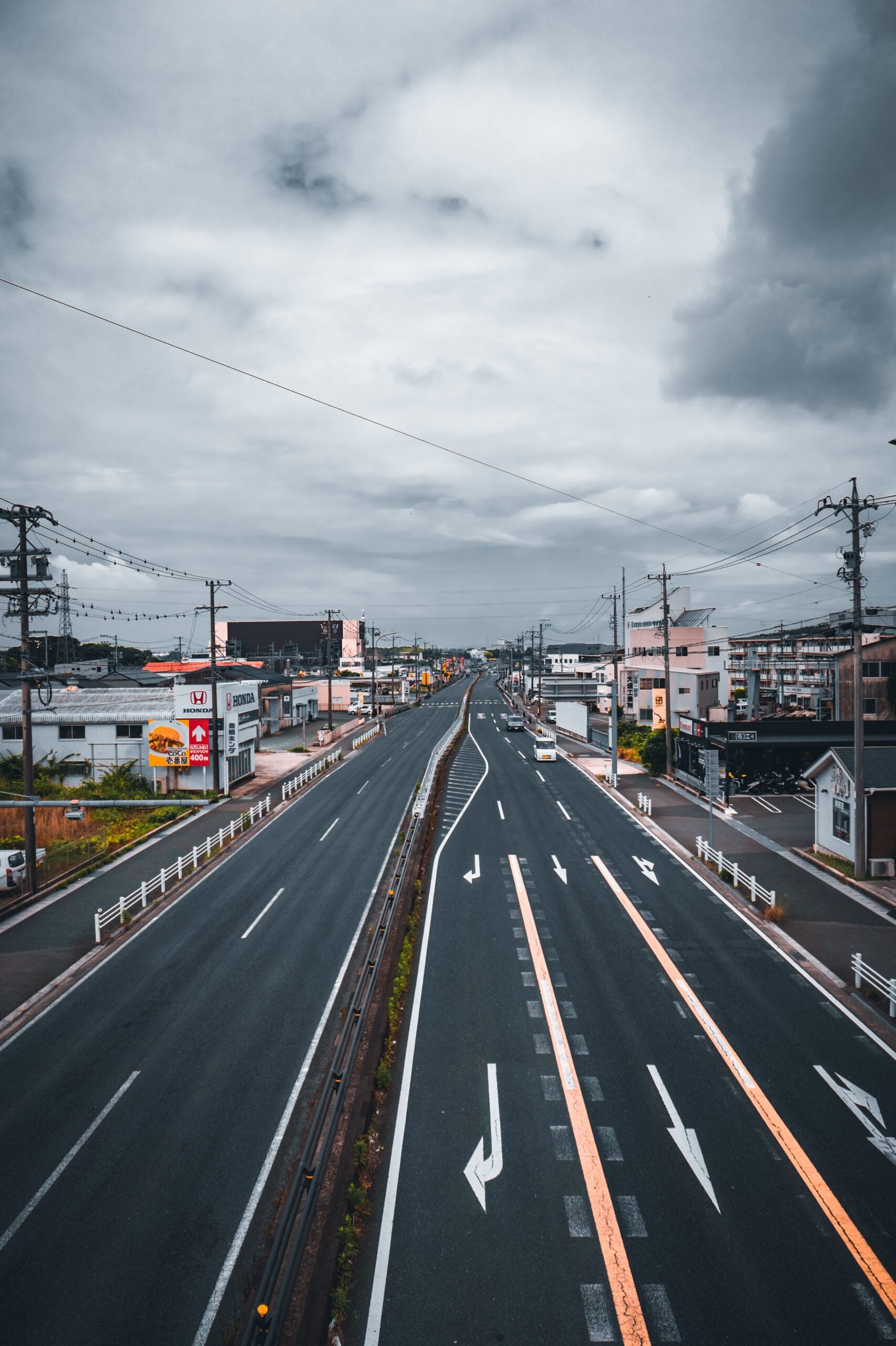 A wide, empty road stretches into the distance between low-rise buildings under a cloudy sky, with lanes marked by white arrows and power lines running alongside.